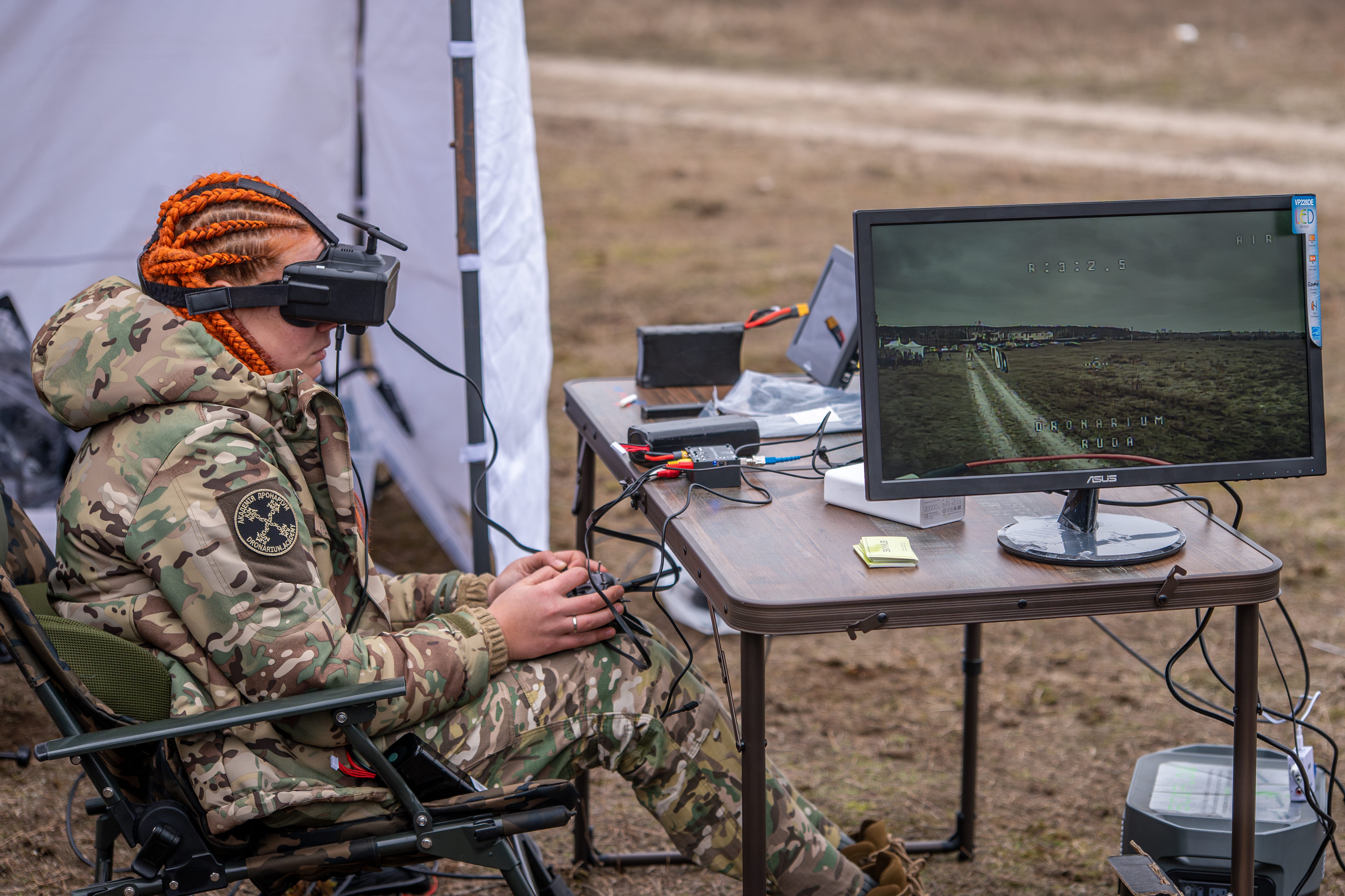 A Ukrainian servicewoman wearing a headset sitting at a desk outside, operating a fiber-optic drone with a remote.