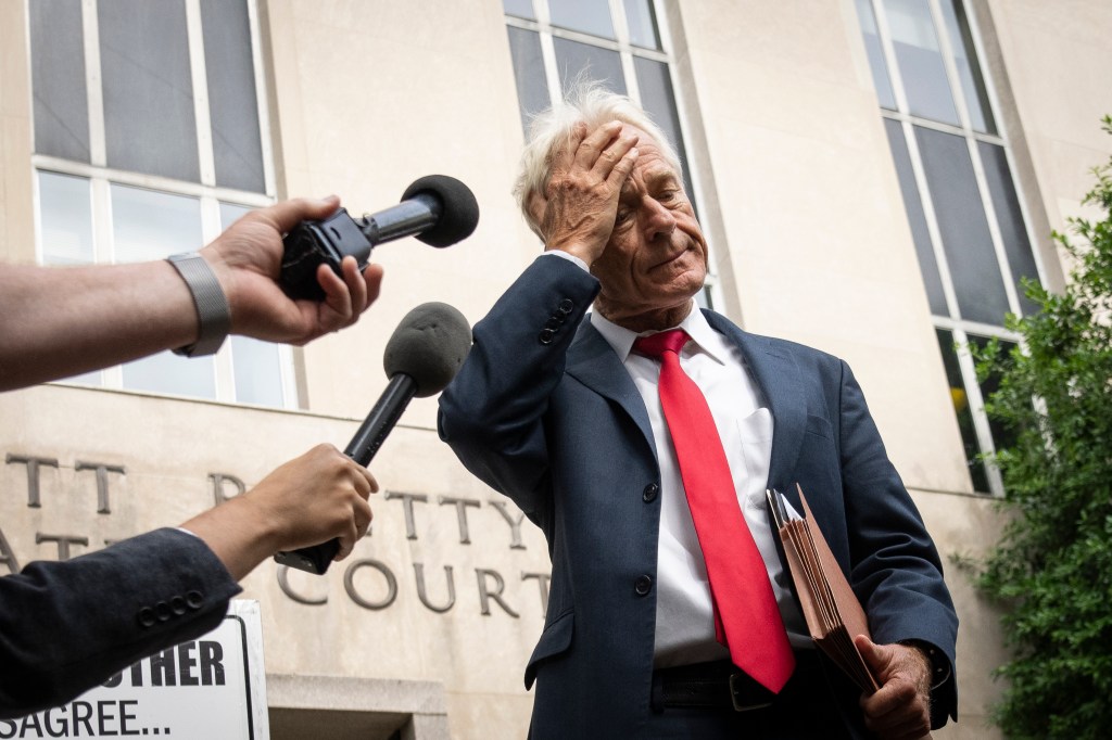 Man in suit holding files, appearing distressed, speaks to reporters with microphones.