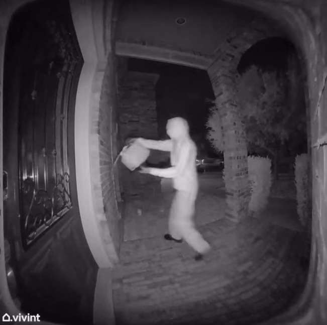 Night vision security camera footage of a hooded person placing a package in a mailbox.