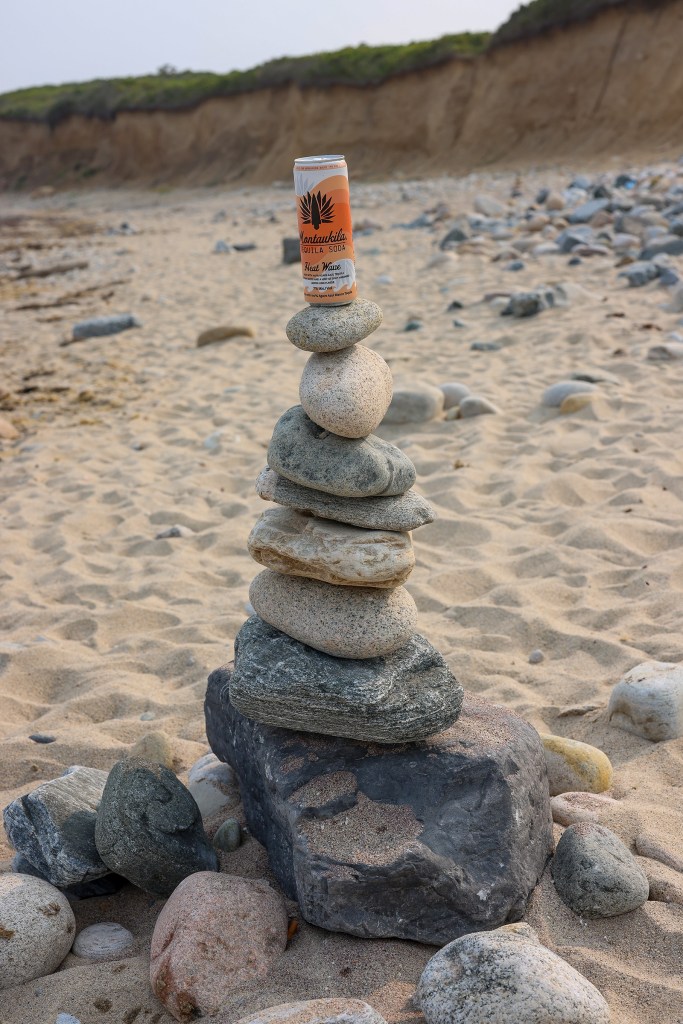 A can of Montaukila Heat Wave tequila soda balanced on a stack of rocks on a sandy beach.