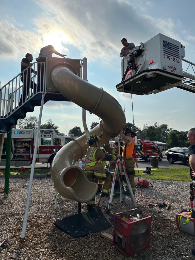 Firefighters using a ladder and a firetruck's extended ladder to remove a child from a playground slide.