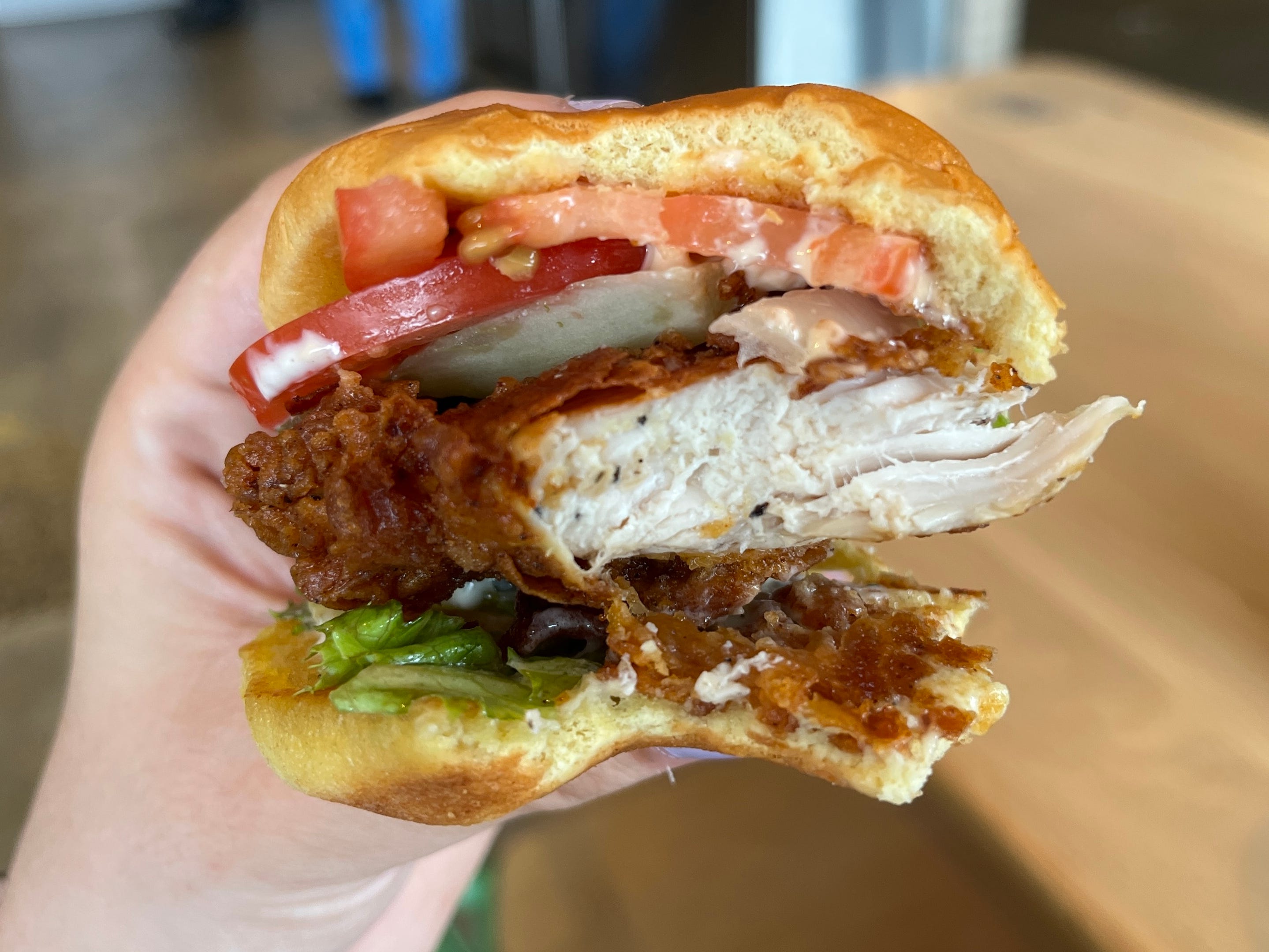 A hand holding a half-eaten fried-chicken sandwich with lettuce, pickles, tomatoes, and onion.