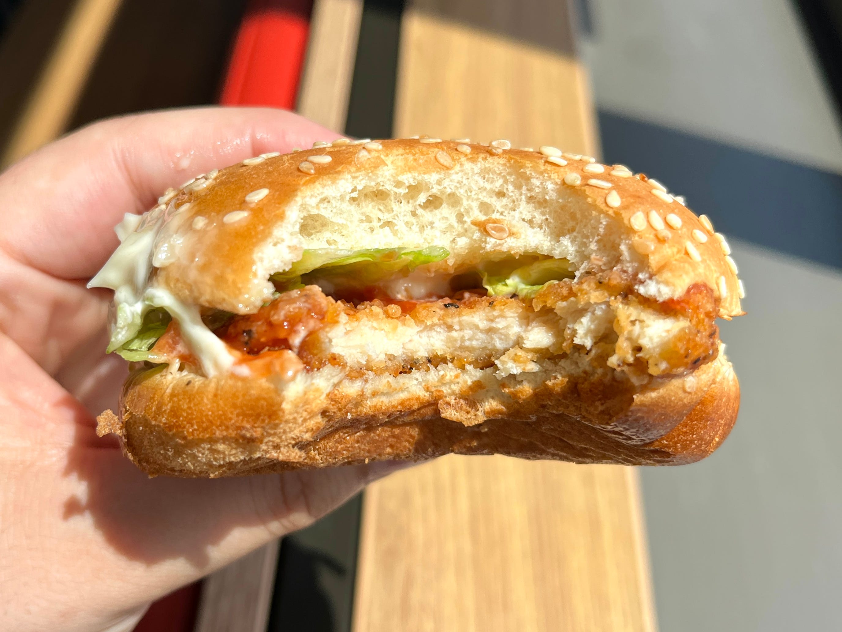 A hand holding a partially eaten fried-chicken sandwich with mayo, ketchup, and lettuce on a sesame-seed bun.