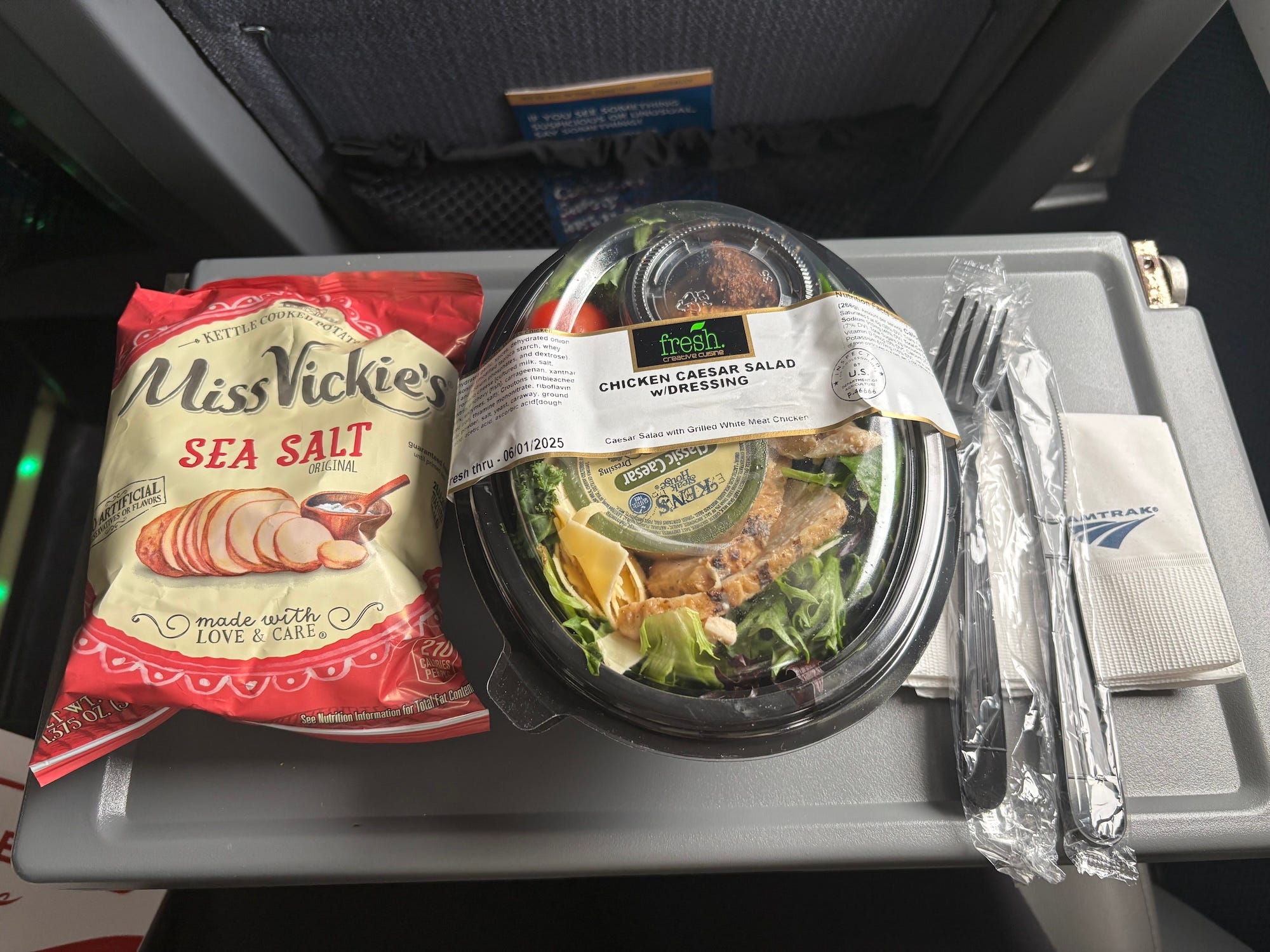 A bag of chips, a pre-packaged caeser salad, plastic silverware, and an Amtrak napkin on a tray table on a train.