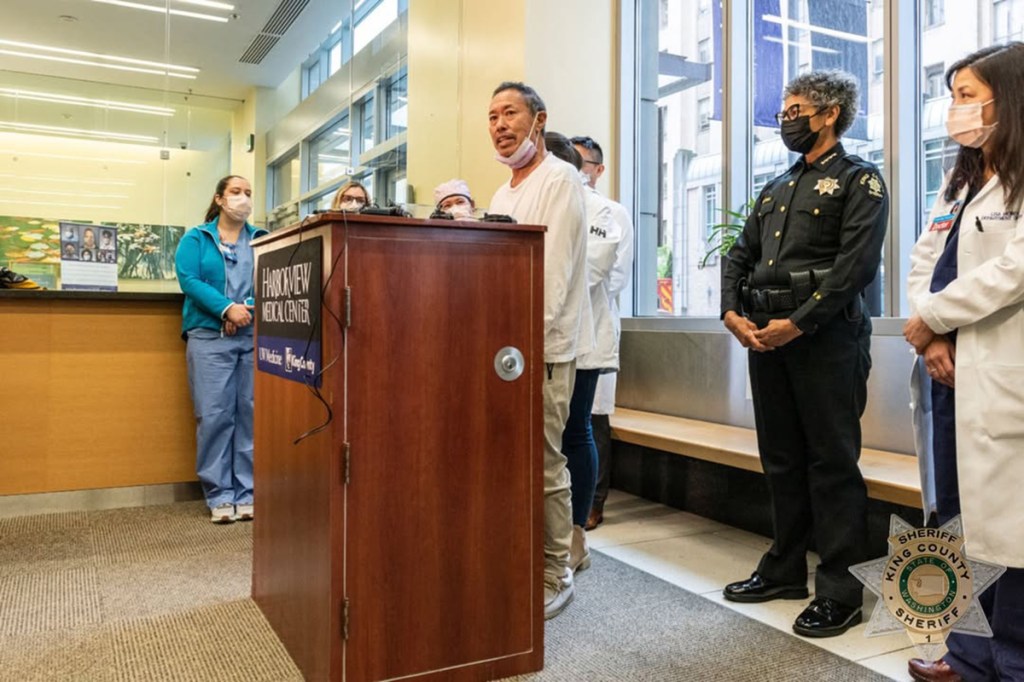 A man speaks at a podium flanked by medical personnel and a sheriff.