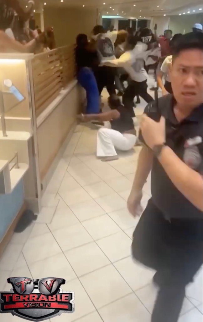 A large group of people fighting in a building; a security guard approaches.