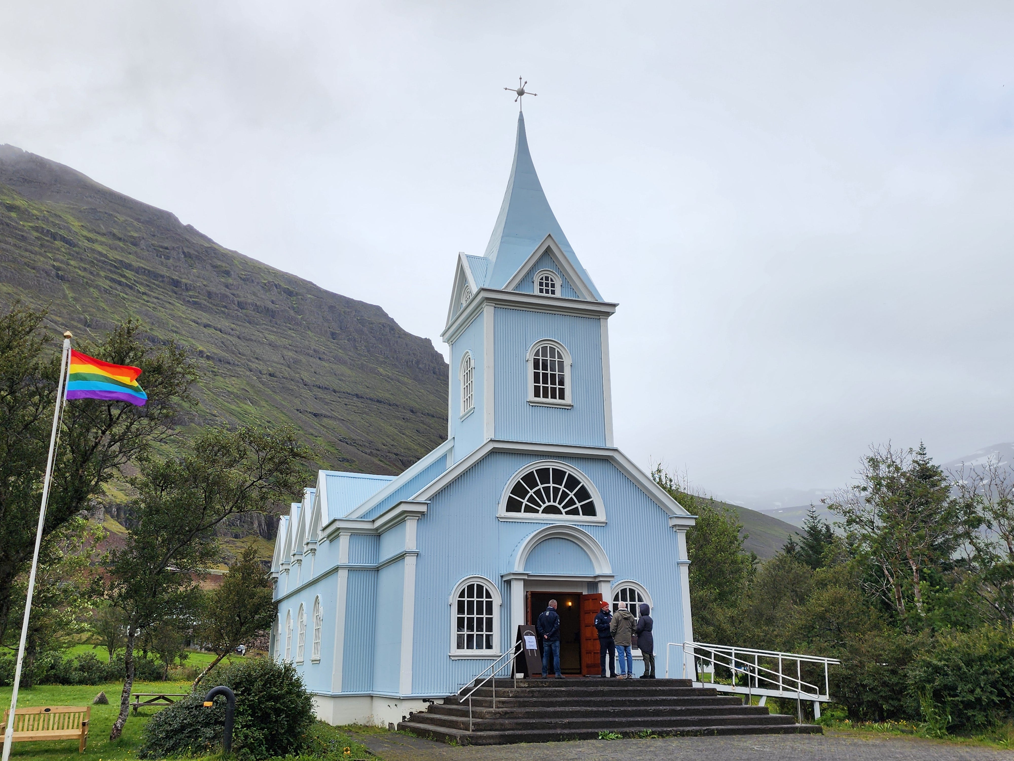 A small blue church in a picturesque countryside setting. It has a rainbow flag flying outside it.