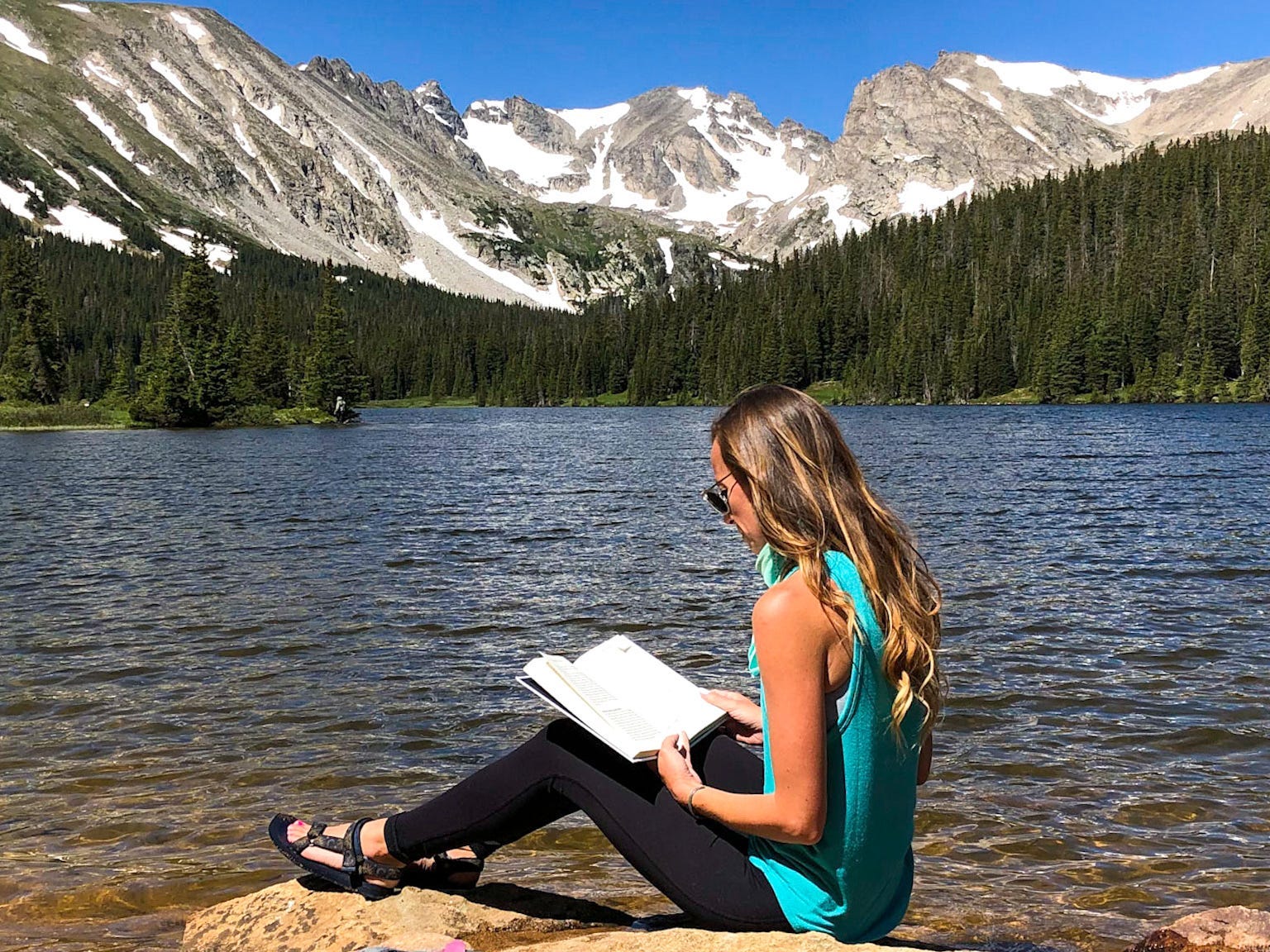 A woman sitting on a rock near a lake, reading a book. The lake is surrounded by trees and the mountains in the background have a dusting of snow.