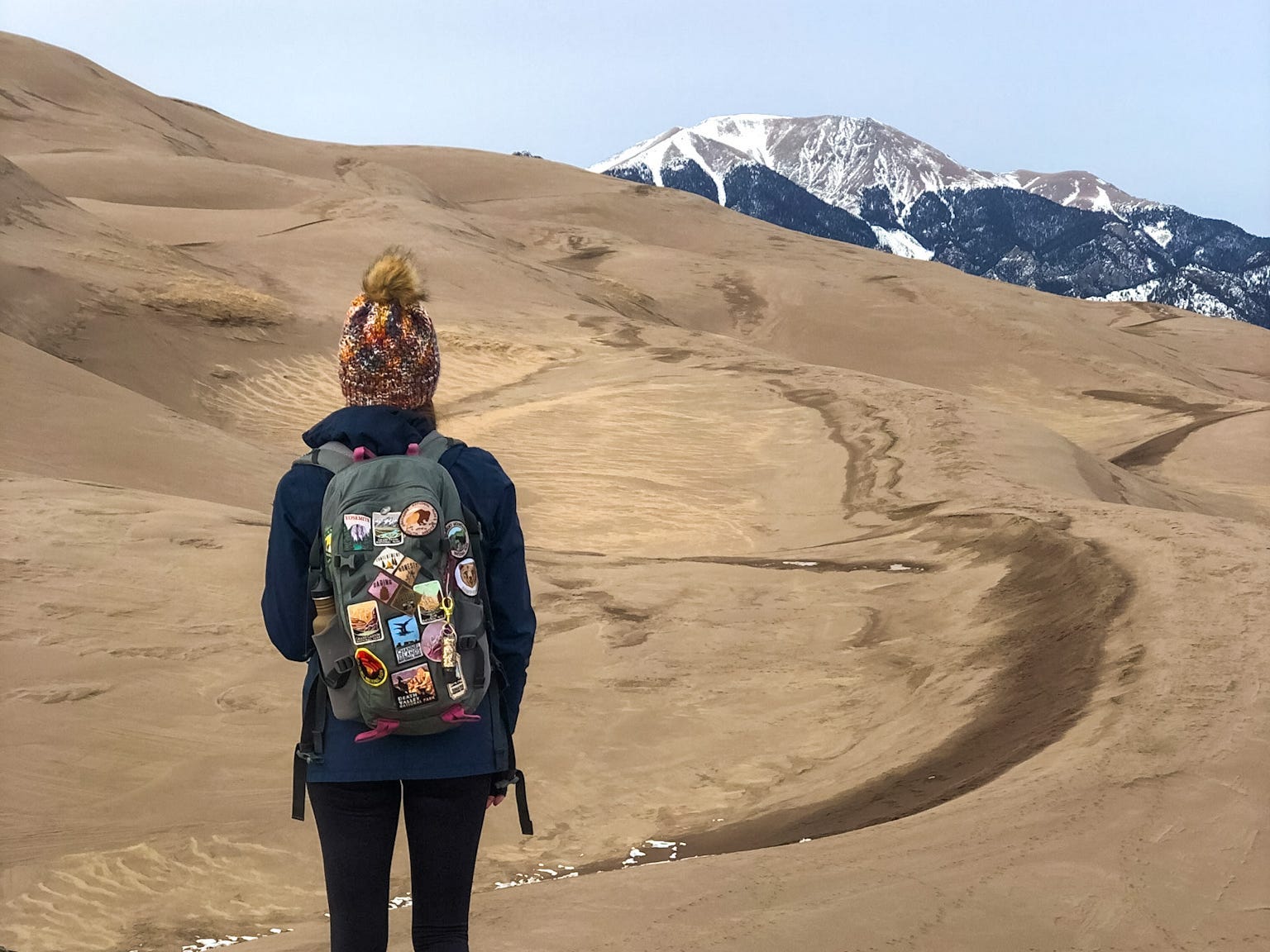 A woman wearing a backpack and wooly hat looking out onto sand dunes with snow-capped mountains in the distance.