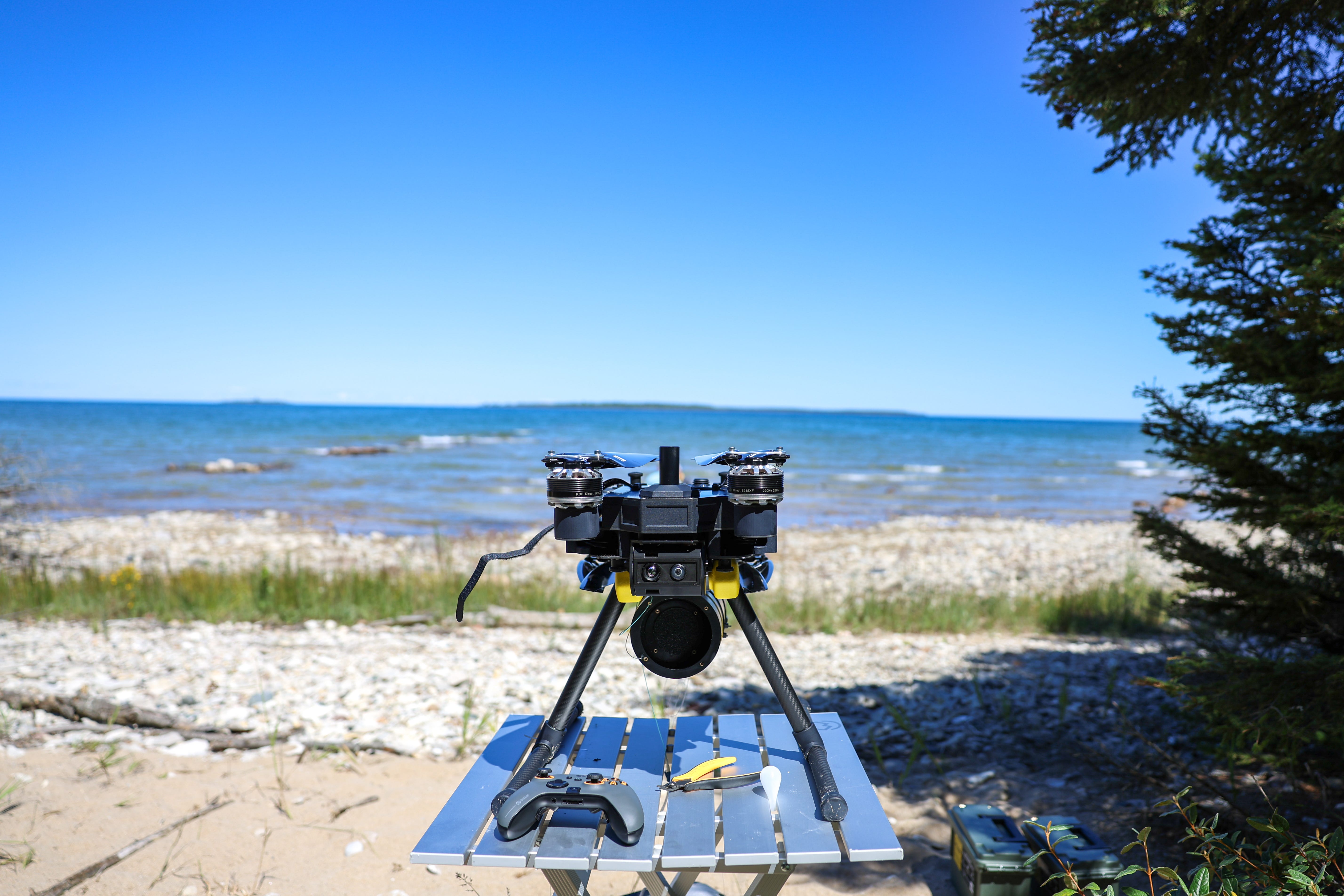 A small, black quadcopter drone sits on a table next to a controller on a beach, with a clear blue sky in the background.