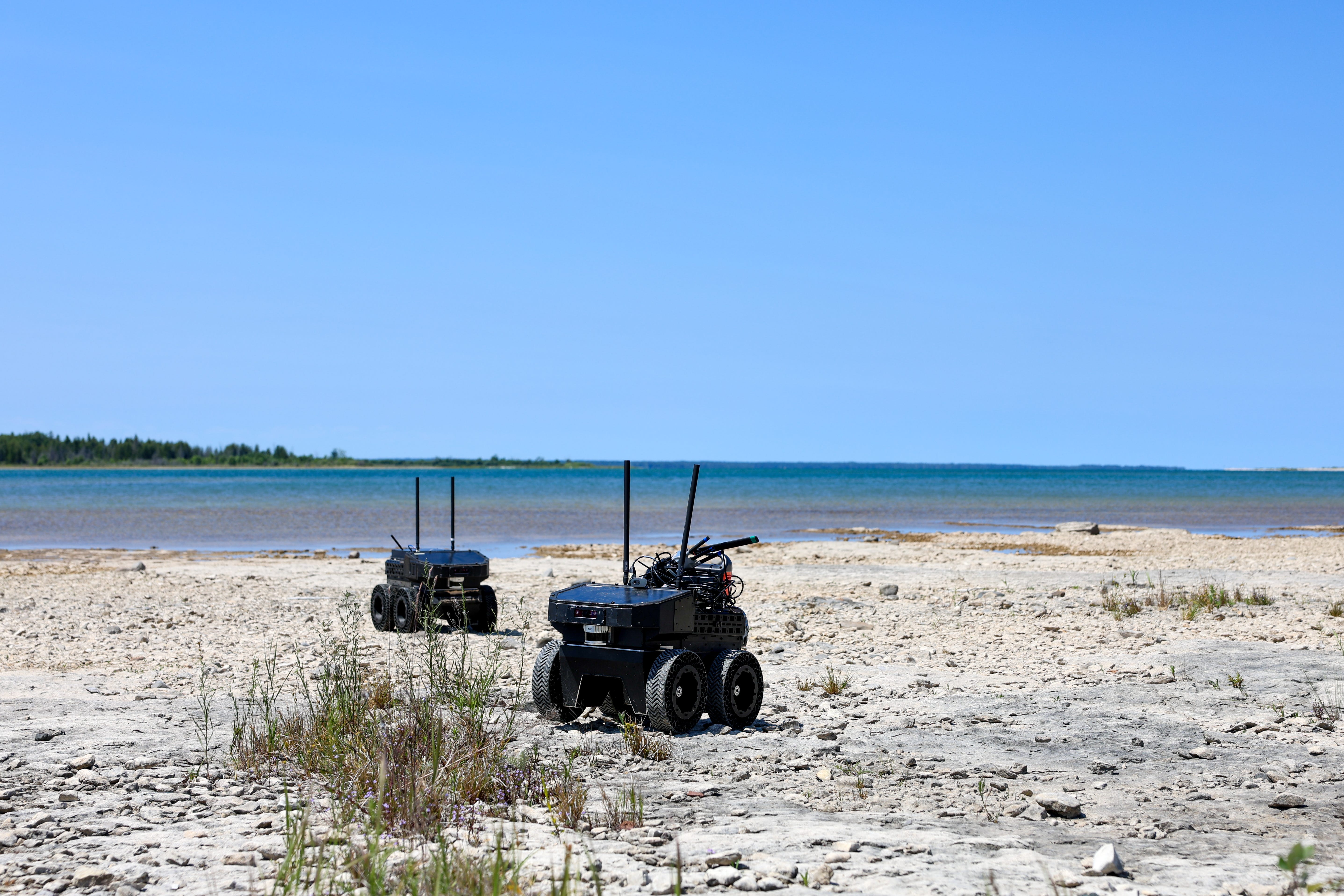 Two small, black ground drones on wheels sit on the beach with a clear blue sky in the background.