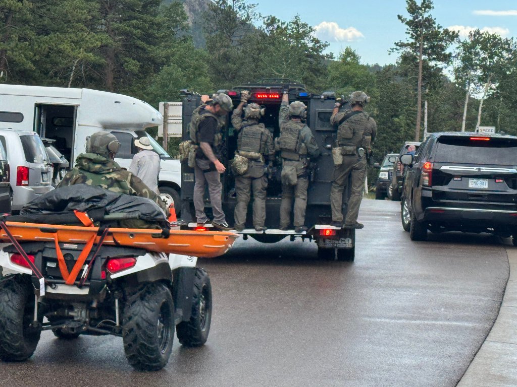 Law enforcement officers in tactical gear on the back of a vehicle at a scene.