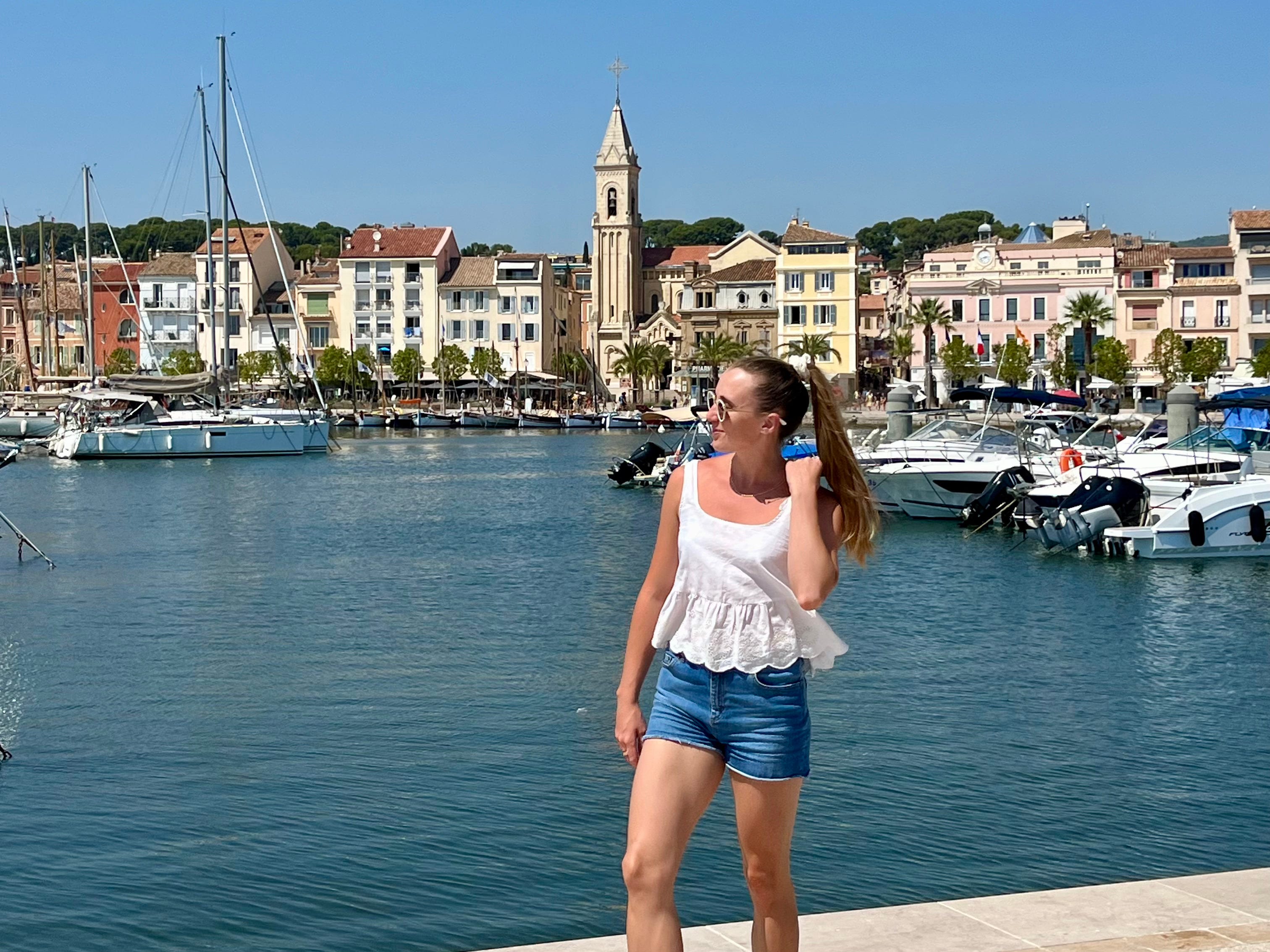 woman in jean shorts and white shirt at a dock with colorful buildings and white boats in the background