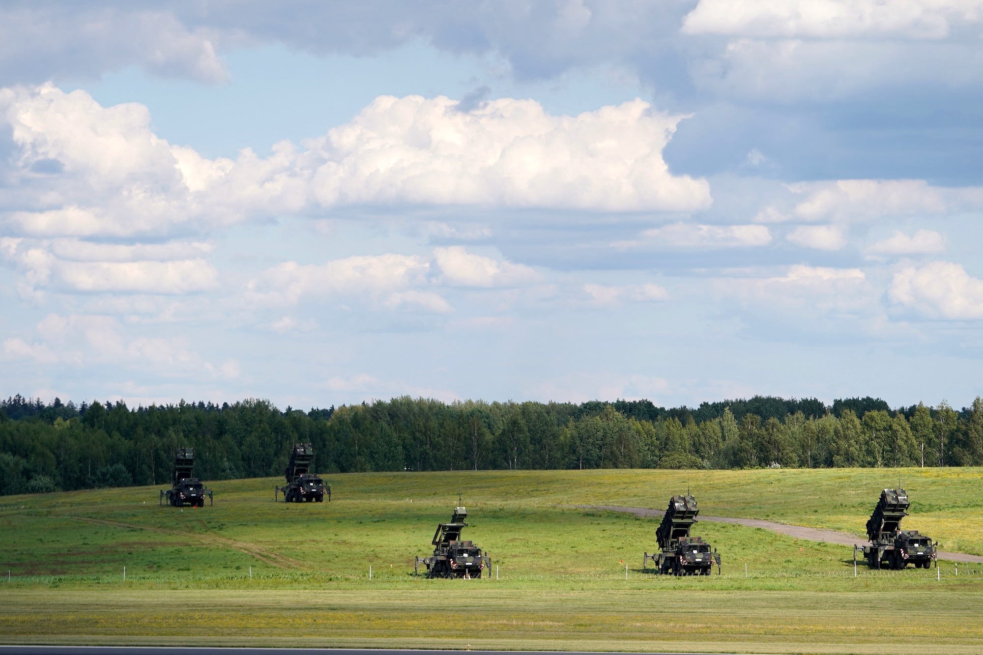 Five Patriot air defense systems sit in a green field under a blue sky
