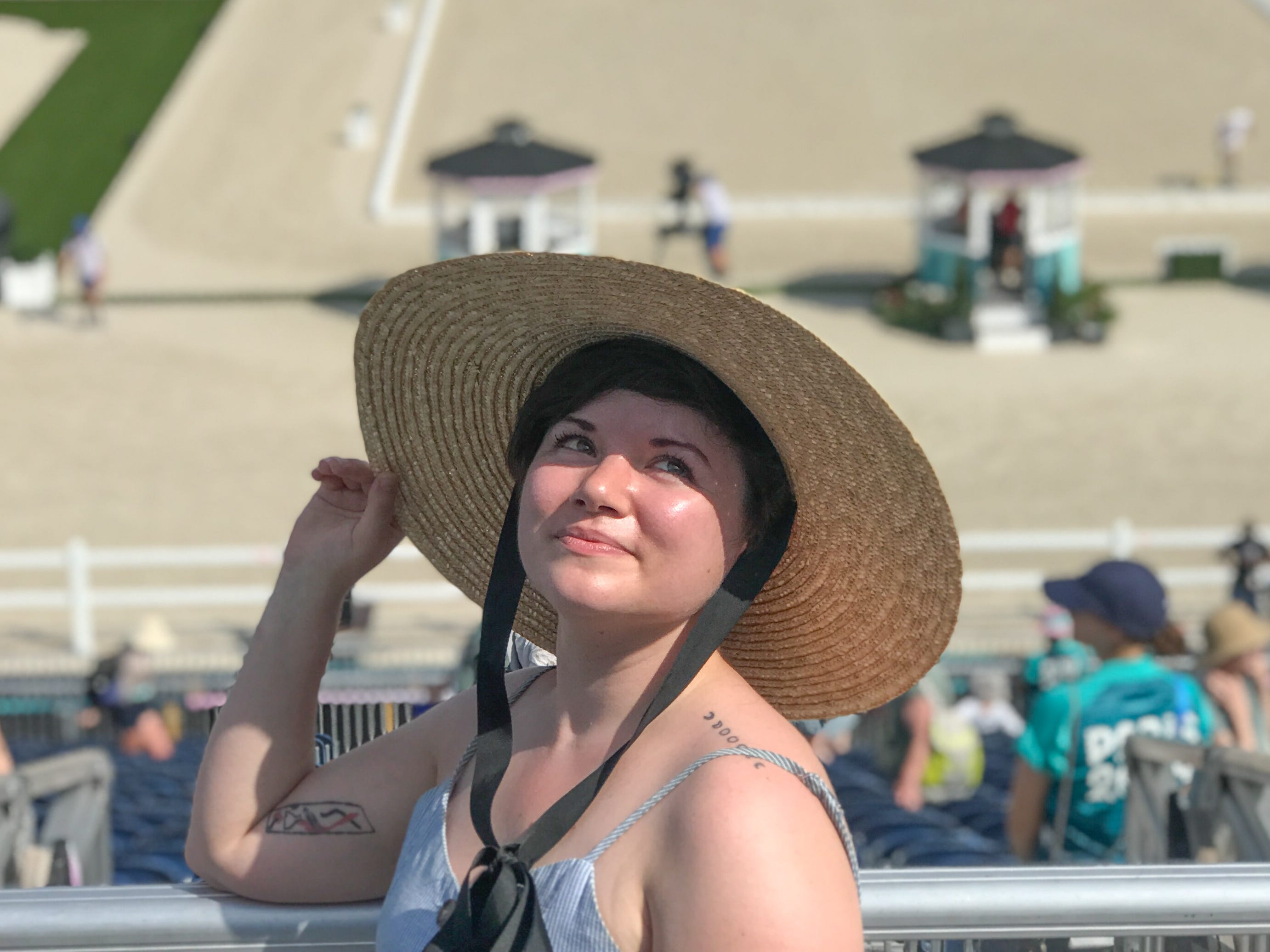 The author smiling at looking up at an event at the 2024 Paris Olympics. She's wearing a wide-brimmed sun hat with a black bow.