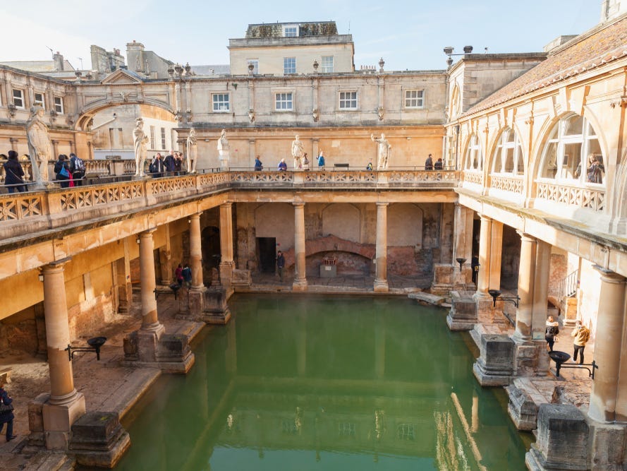 Tourists walk around a two-floor open-air Roman bath. The bath in the middle of the building is green.
