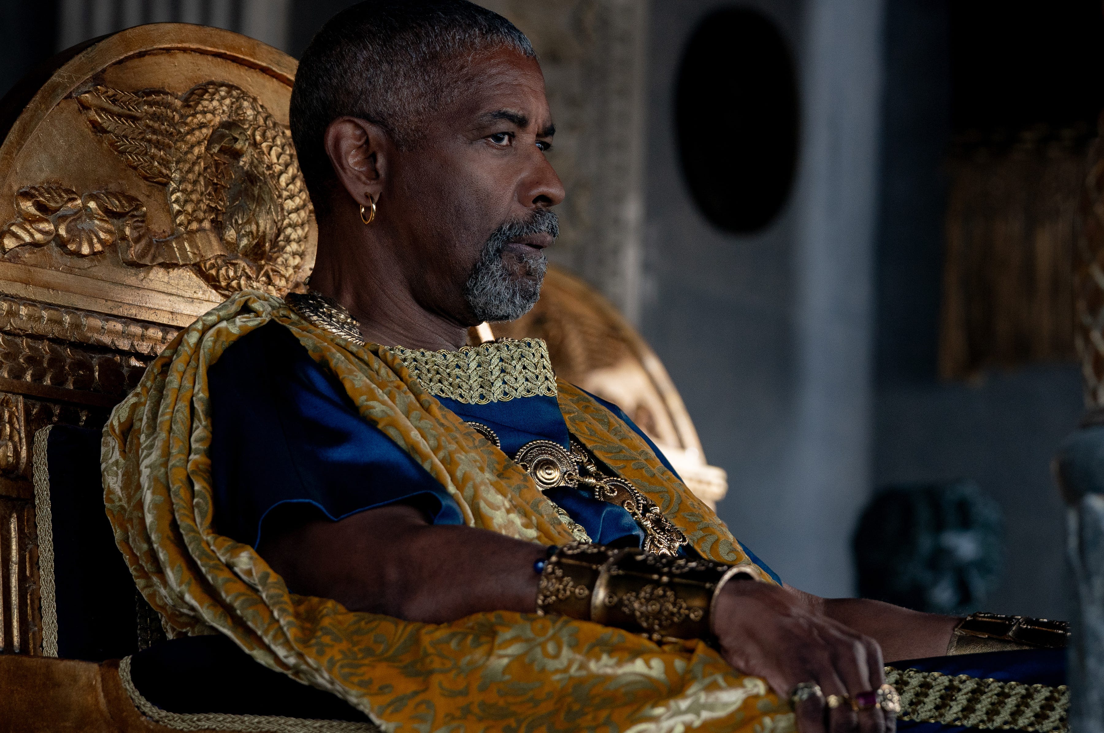 denzel washington as macrinus in gladiator two. he's wearing ornate blue and gold robes and has on rings, bangles, and earrings, and looks skeptically outwards from his place on an ornate chair