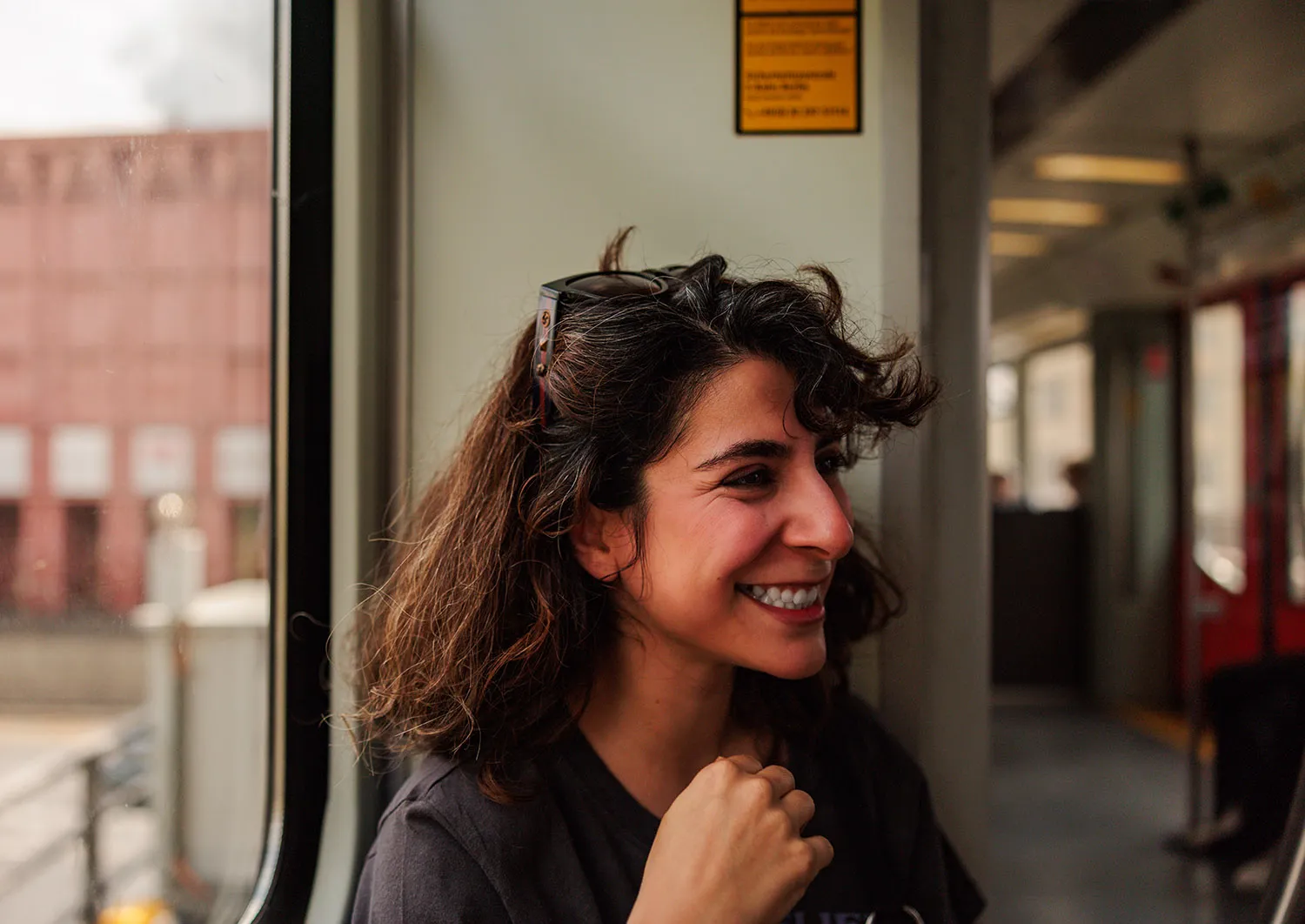 A woman with sunglasses on her head smiles and looks to the side as she sits next to the window of a train.