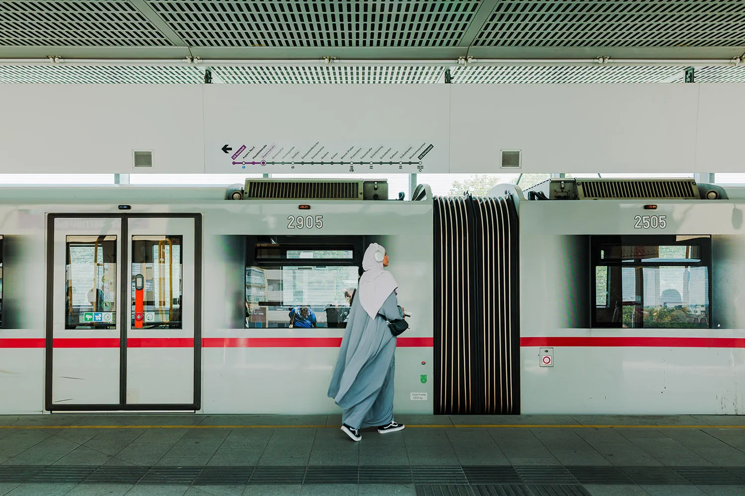 A woman in a headscarf and long flowing garment, tennis shoes, and large headphones steps toward a white train on a platform.