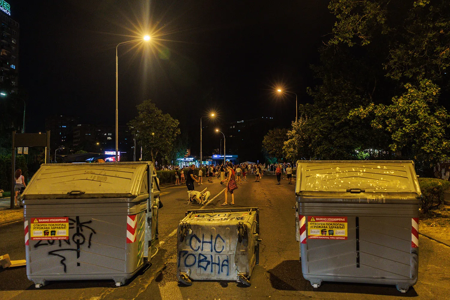 Large trash bins block a street as people are seen in the distance as streetlights light up the night scene.