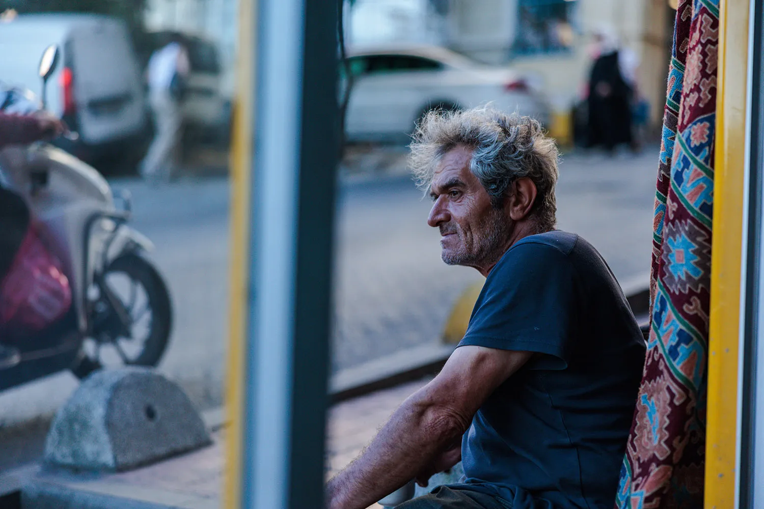 A man with gray hair, seen from the side, outside a shop with a colorful rug hung up behind him and an out-of-focus street and vehicles in front of him.
