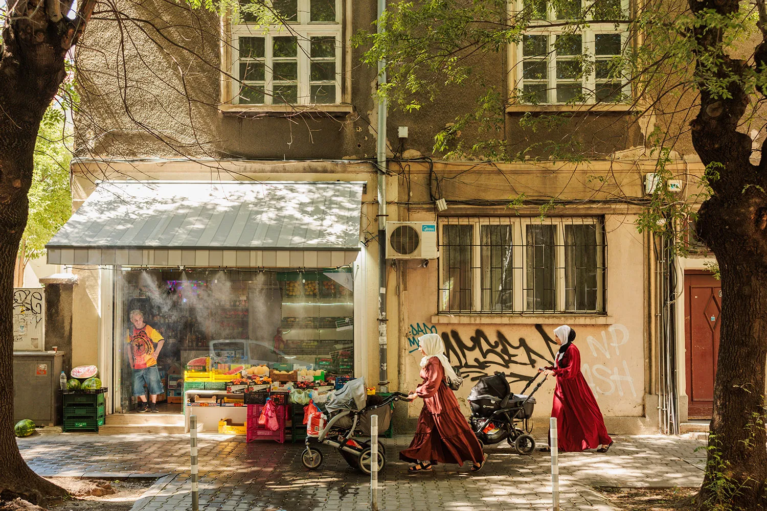 Two women in long red dresses and head coverings push strollers down a street. A shopkeeper stands in the doorway of his shop where misters water fruit out front.