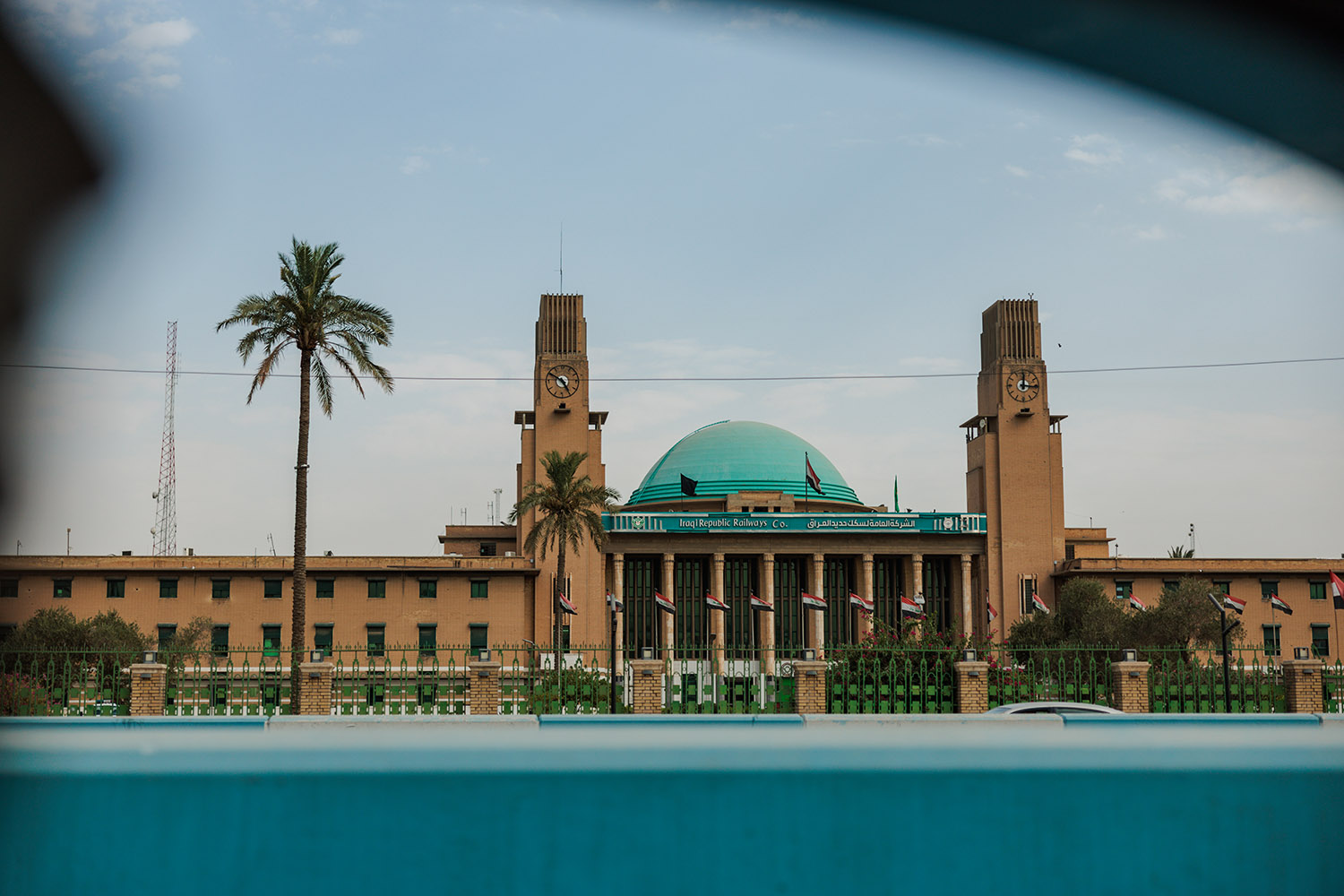 A train station with two clock towers on either side of a turquoise dome, with two tall palm trees in front of it.