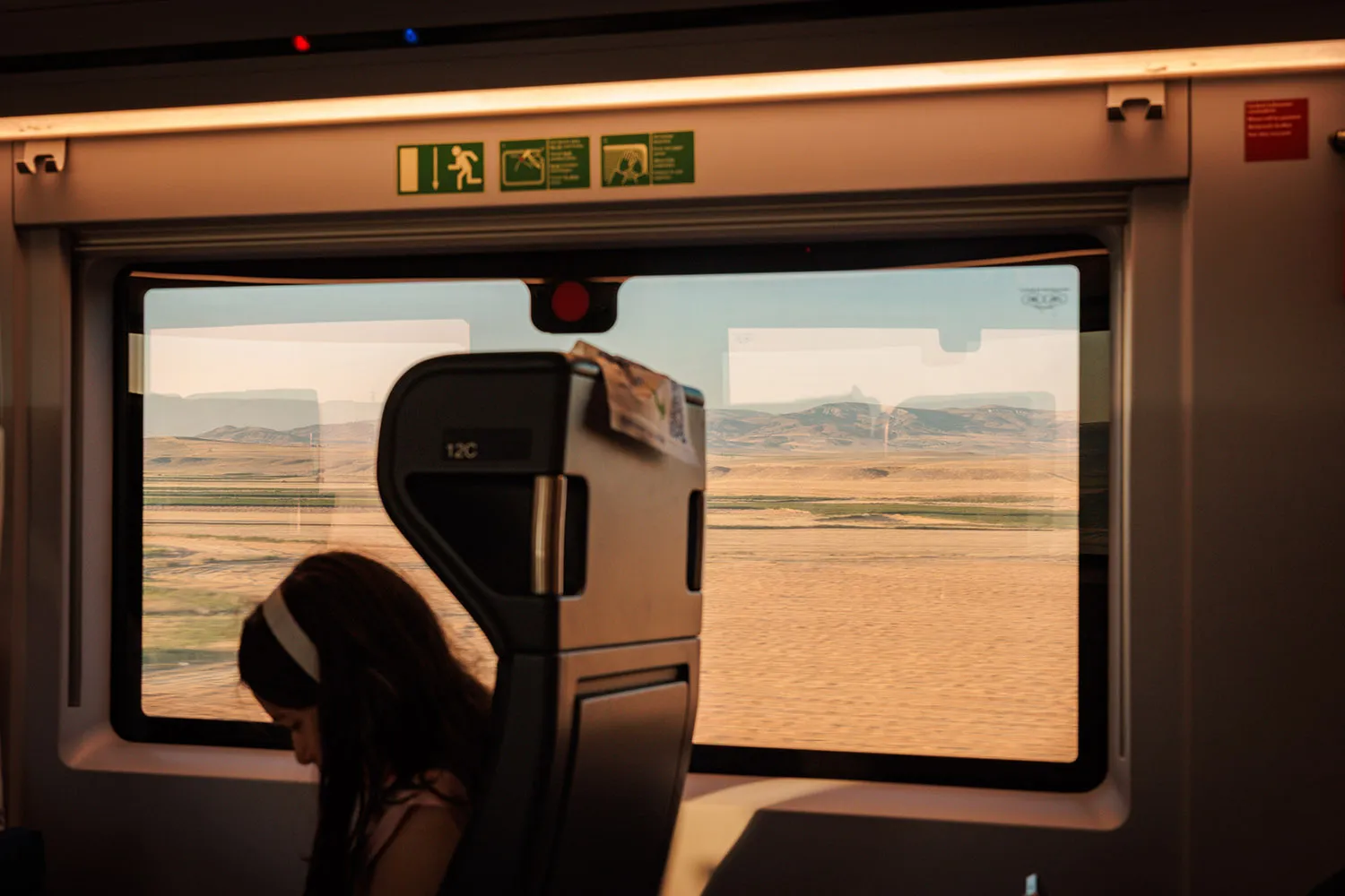 A girl with a headband sits on a train with a desert landscape seen through the large train window.