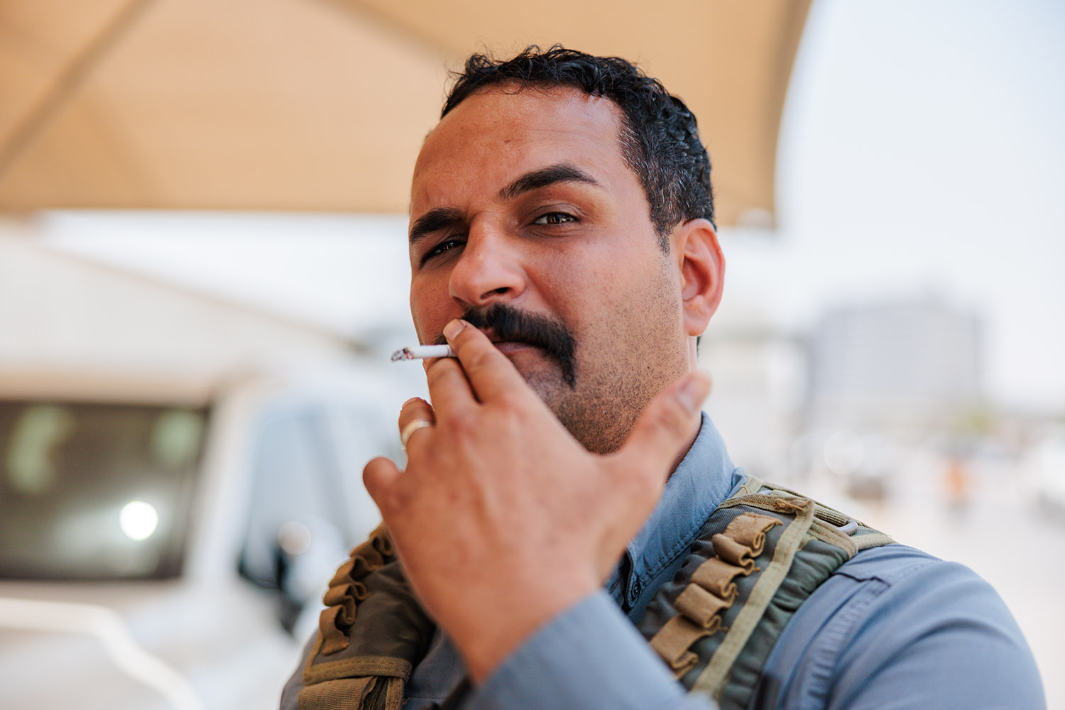 A man squints and lifts one eyebrow as he smokes a cigarette and looks into the camera.