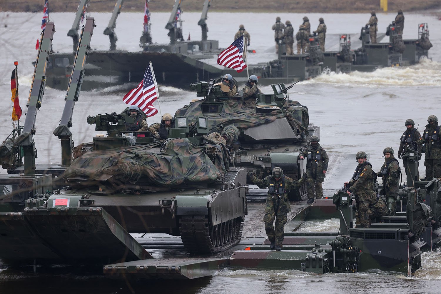 Tanks with U.S. flags on them float on rigs in a river while soldiers accompany them.