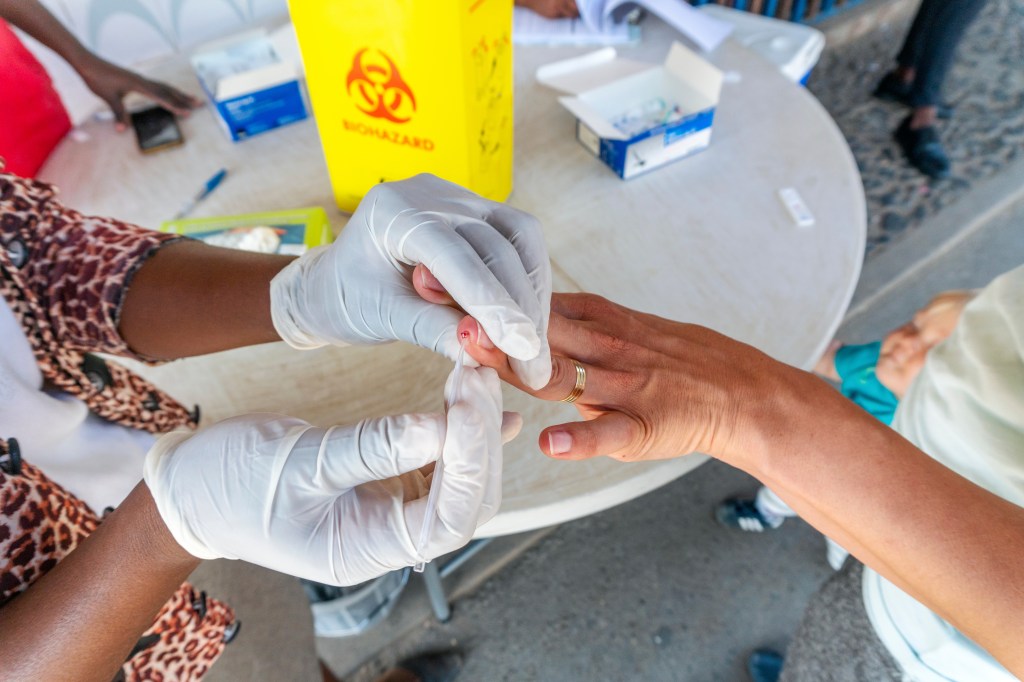 gloved hands pricking someone's finger with a yellow biohazard sign in the background