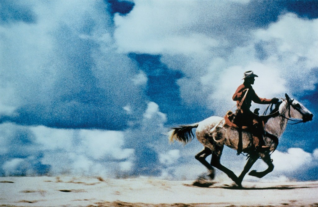Photograph of a cowboy riding a horse across a desert landscape.