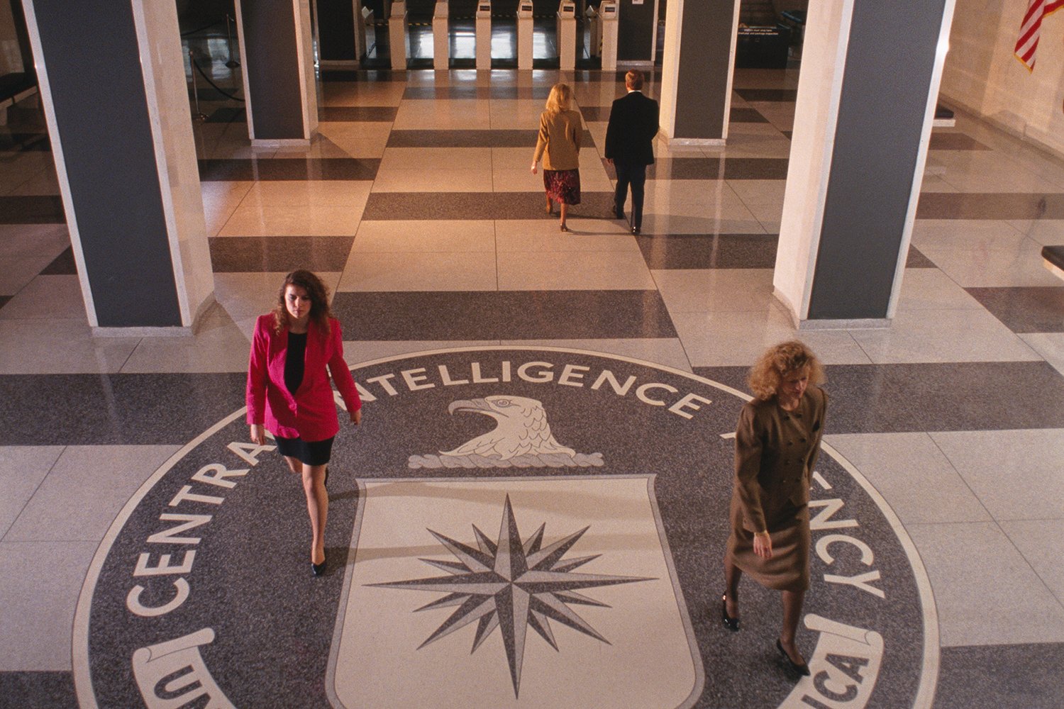 Background is inside a building entrance lobby. In the foreground two women in dress coats and skirts walk either side of the circular Central Intelligence Agency crest placed in the marble floor.