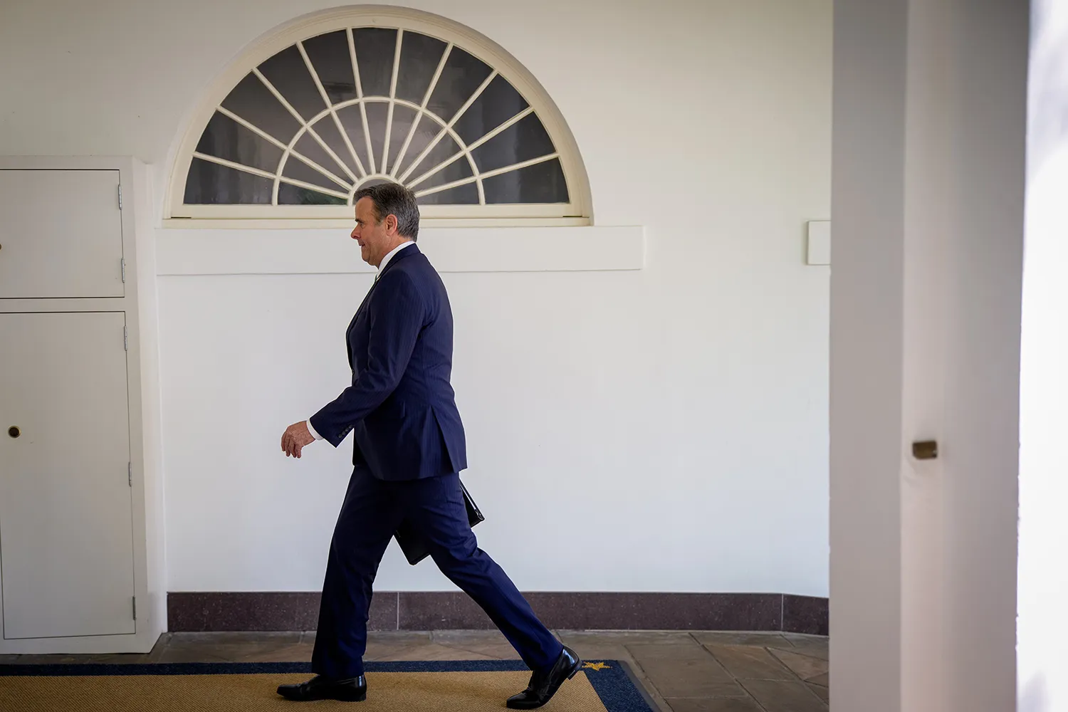 Ratcliffe in a suit walks in profile past a semi-circle window in a white wall.