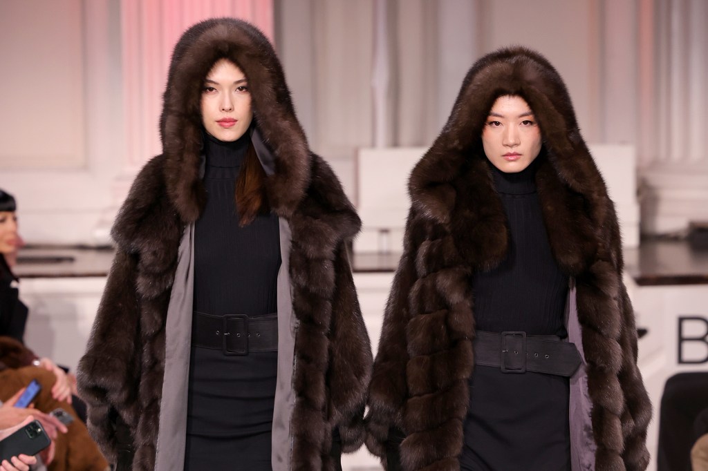 Two models in brown fur coats walk a runway.