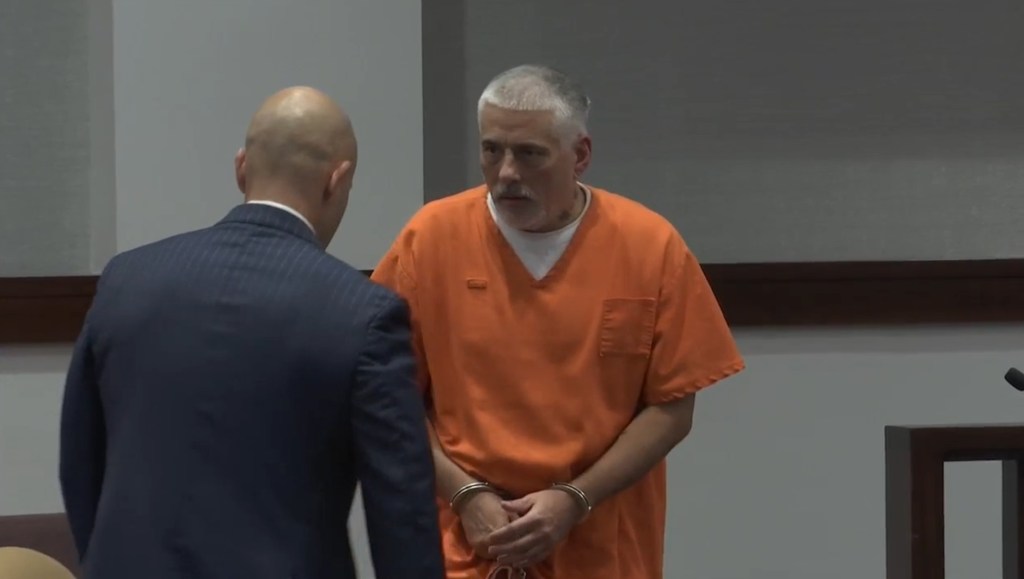 A handcuffed man in an orange jumpsuit speaks with a person in a suit.