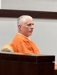 Man in orange jumpsuit seated in courtroom.
