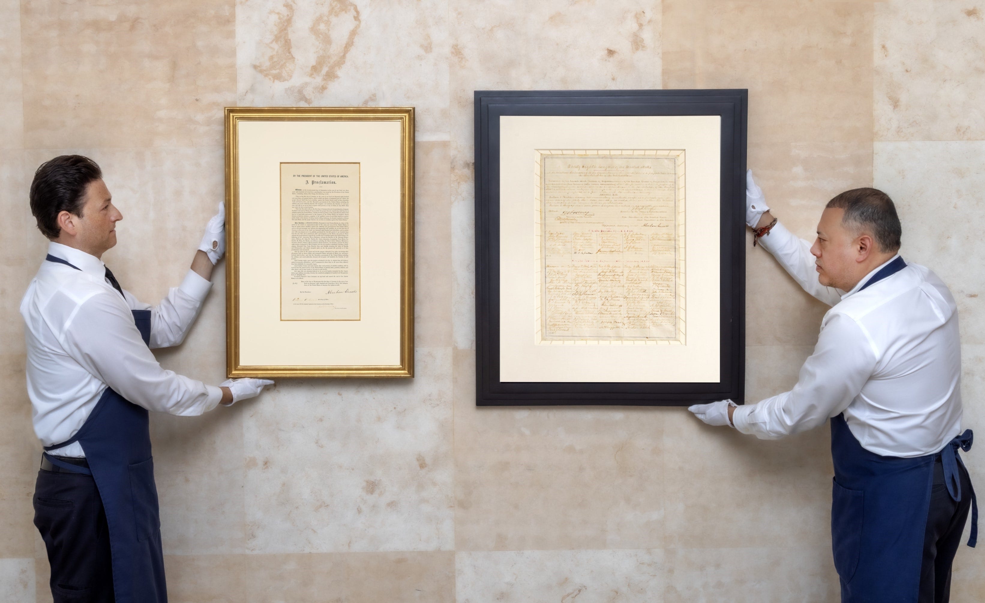 Two men hold up framed documents with their white gloves; the gold frame on the left is a copy of the Emancipation Proclamation, and on the right is the Thirteenth Amendment.
