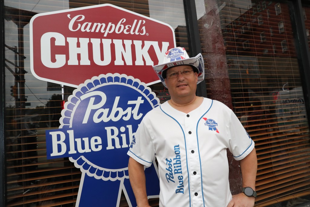 Man in Pabst Blue Ribbon shirt and hat standing in front of a Campbell's Chunky soup sign.