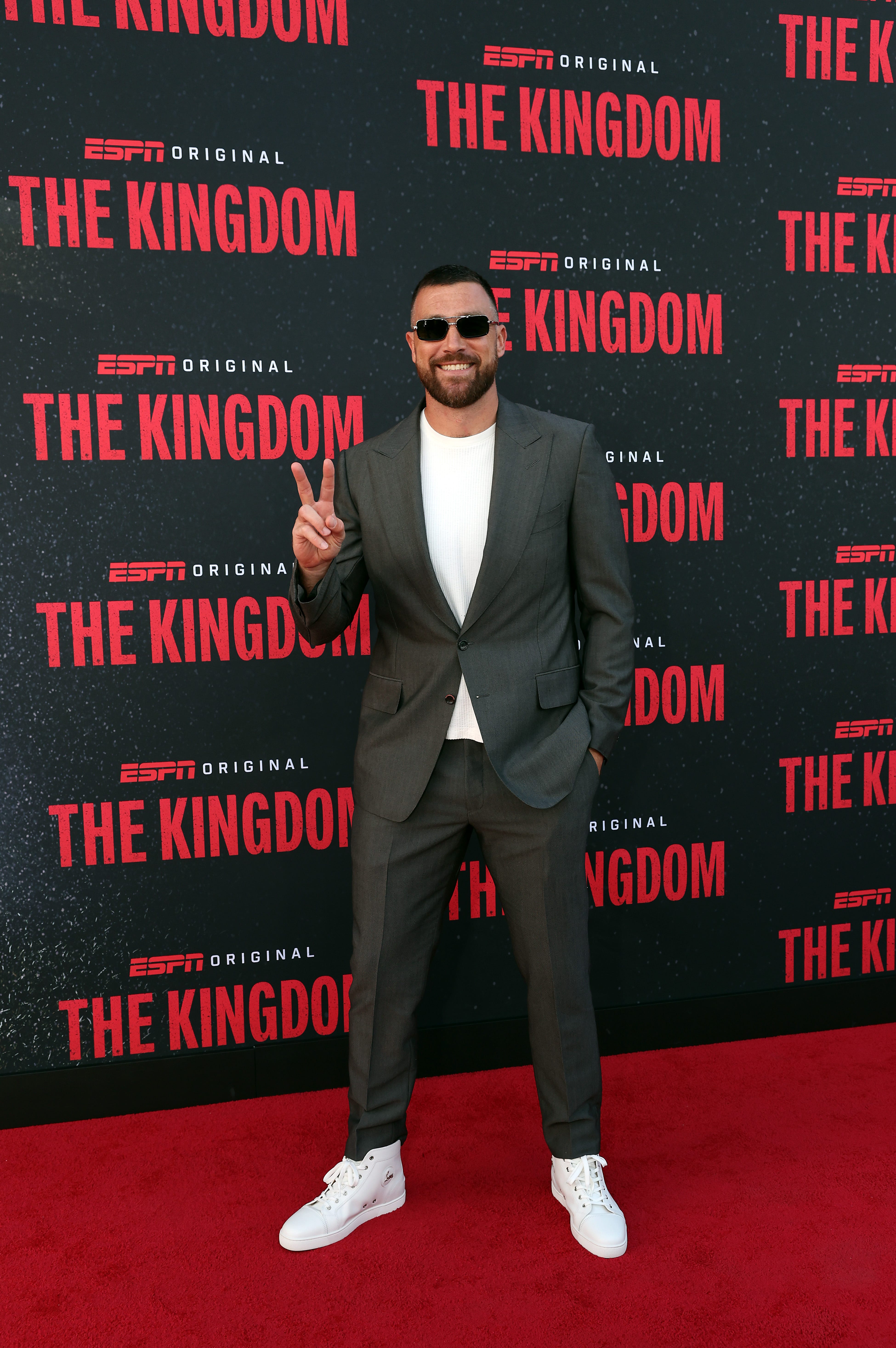 Travis Kelce wears a gray suit and gives a peace sign at a red-carpet screening of "The Kingdom."