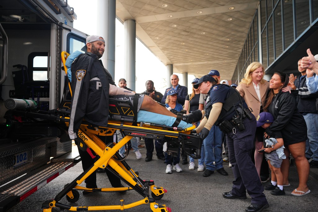 NYPD detective being released from hospital on stretcher, surrounded by family and police officers.