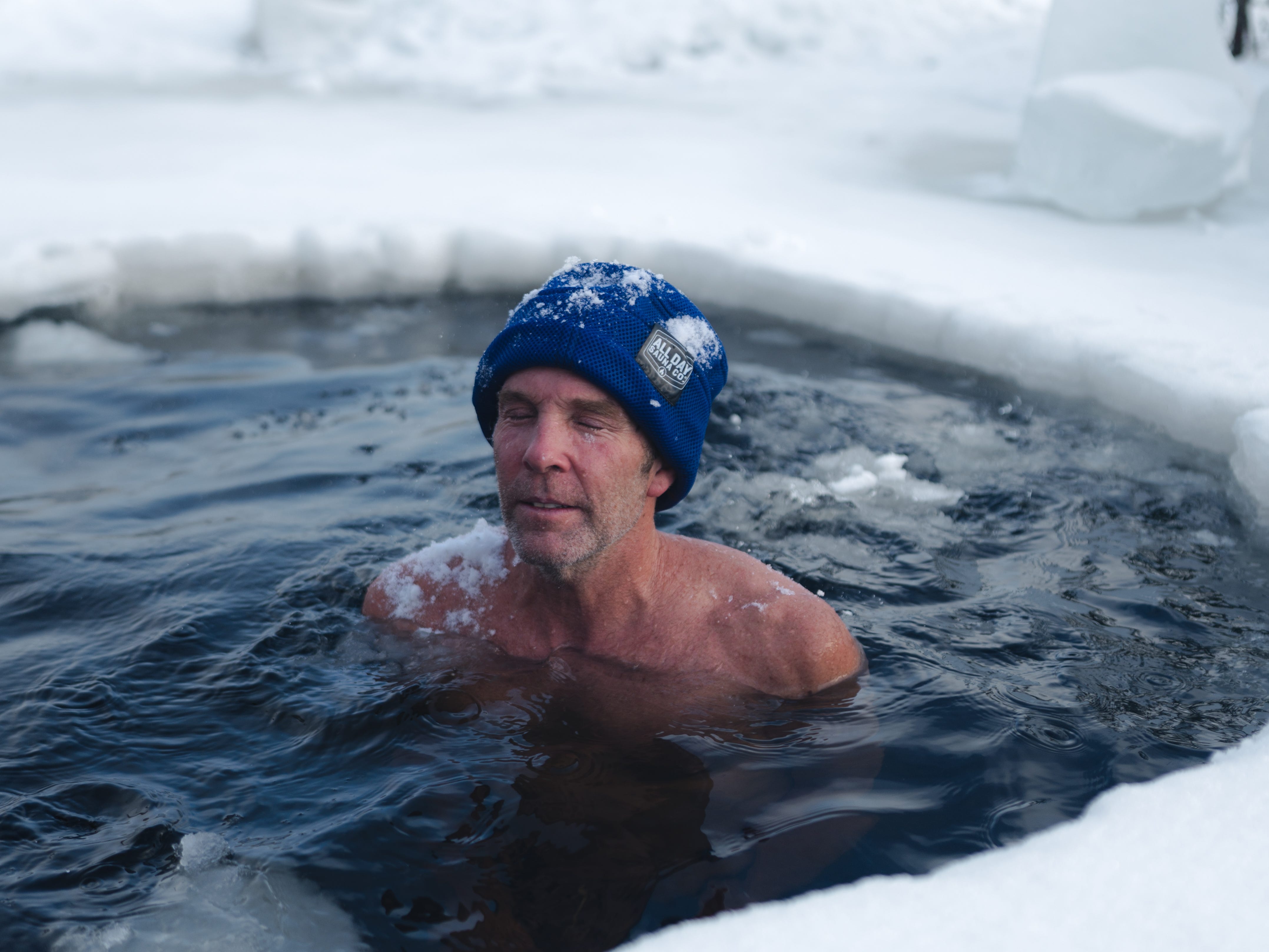 a man in a blue hat doing a cold plunge outdoors surrounded by snow