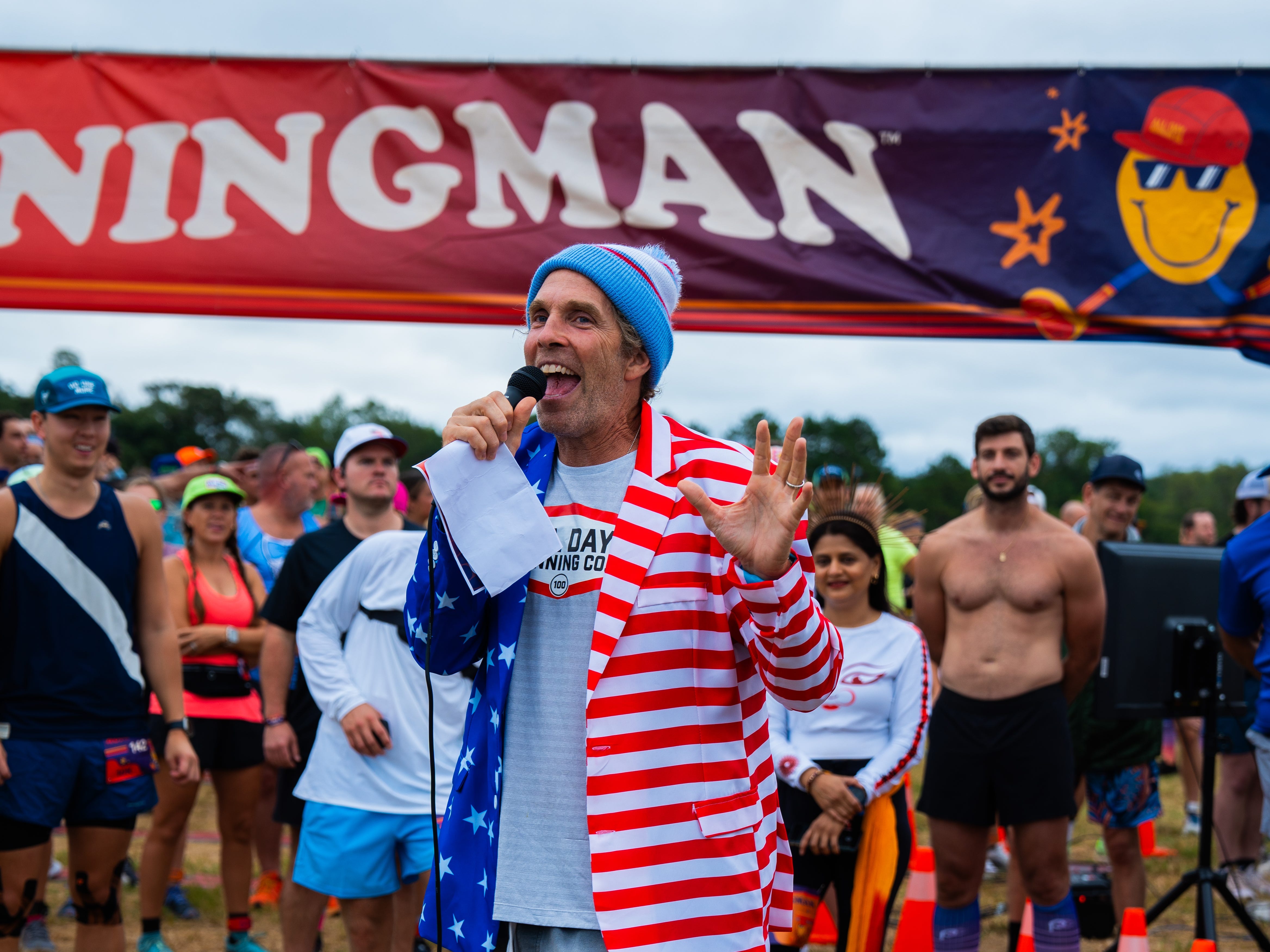 A man in a american flag blazer, running shorts and shoes, speaking into a microphone outside