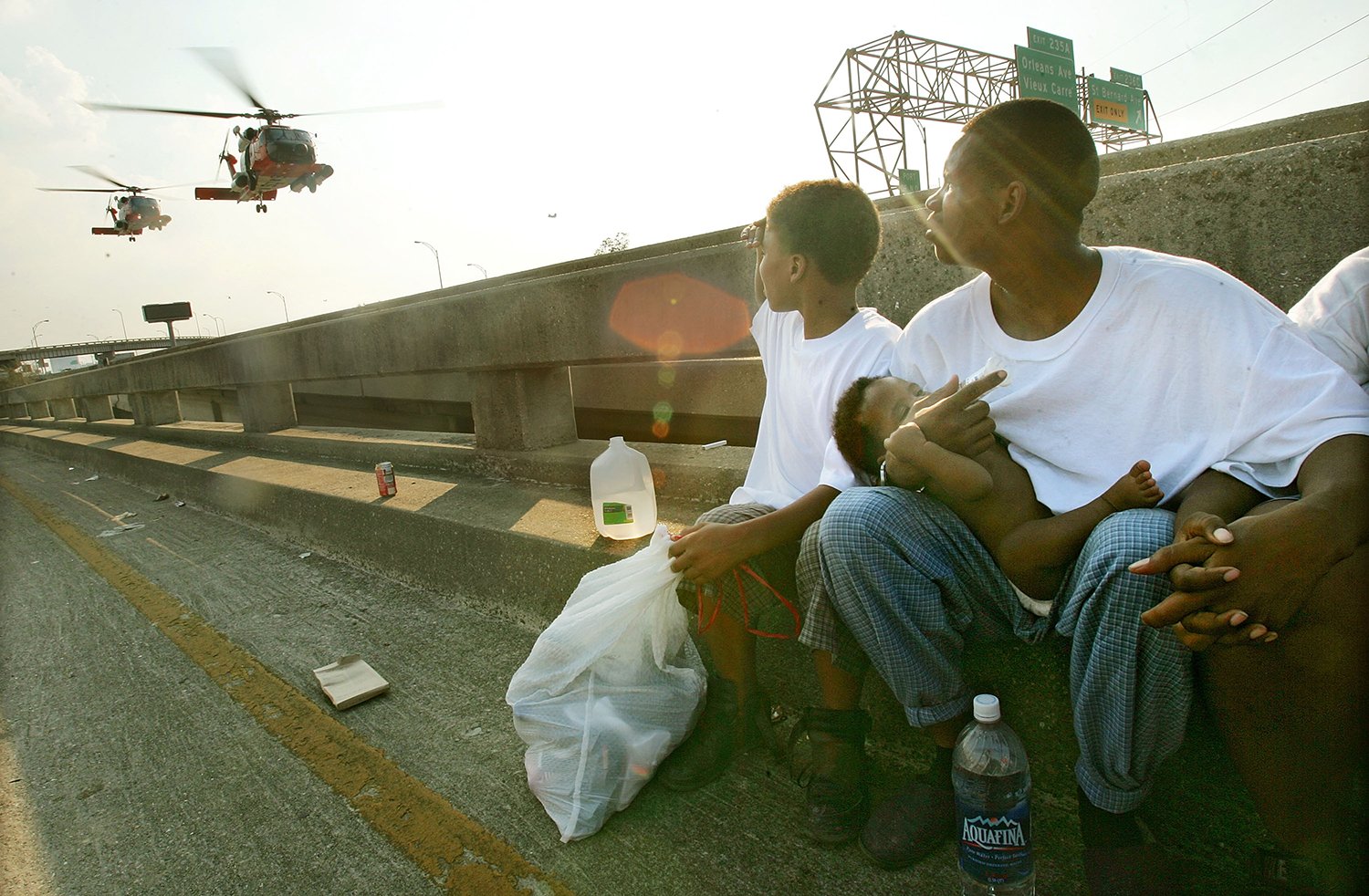 Two young men, one holding a baby, sit on a highway overpass as helicopters fly overhead.