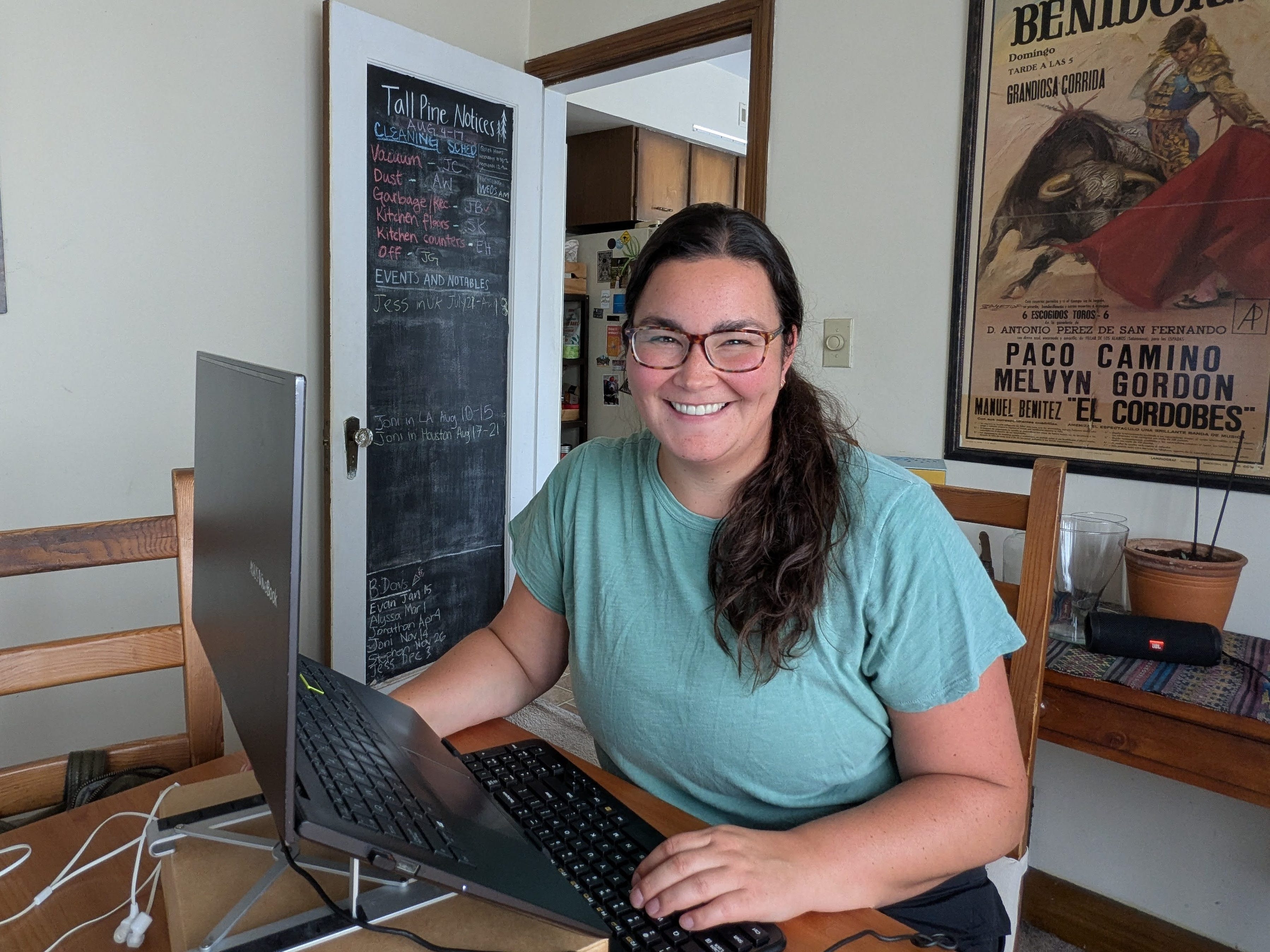 The author smiling from her workspace at the kitchen table. There's a chalkboard door behind her and artwork.