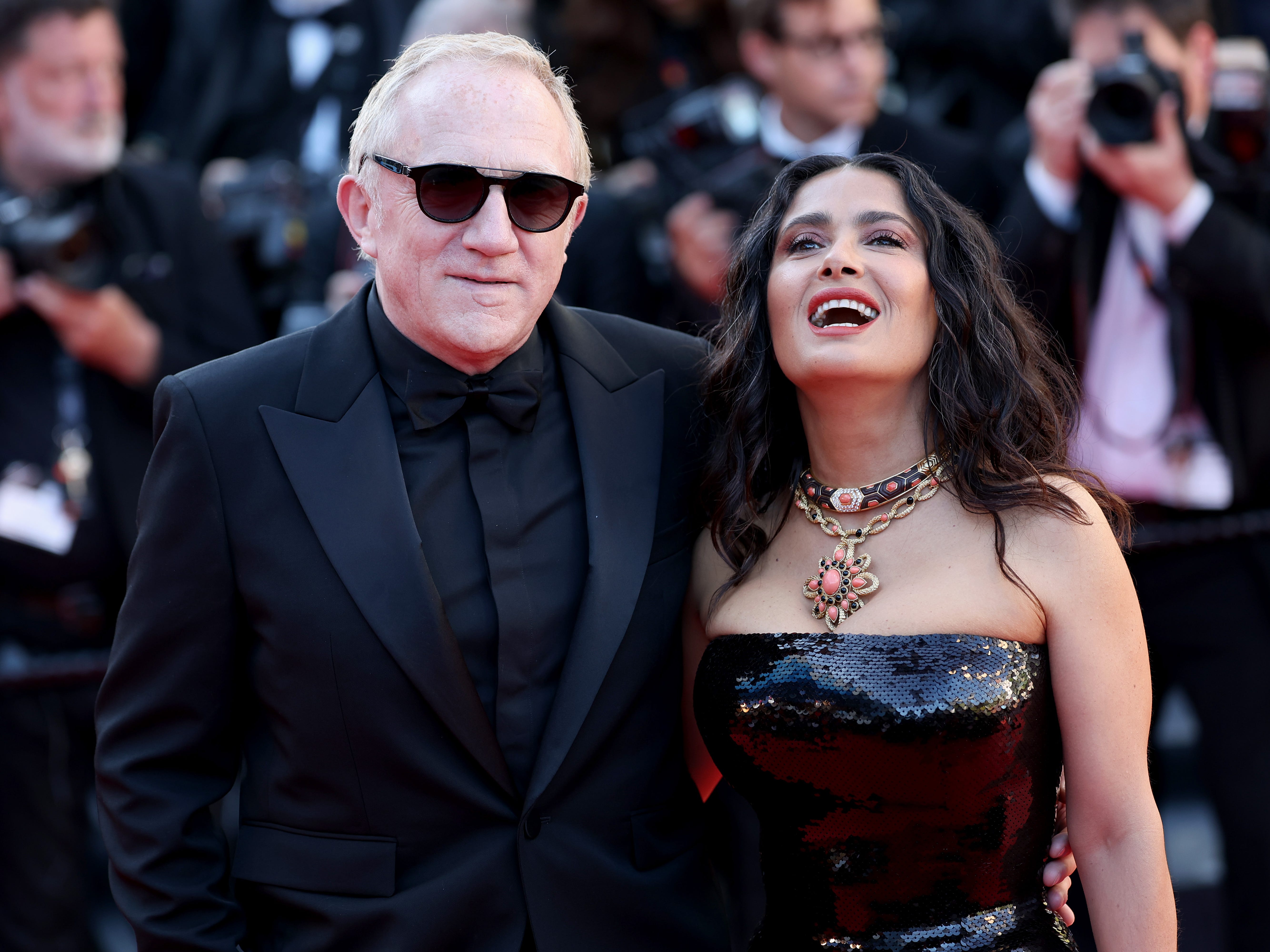 François-Henri Pinault, wearing a black suit and shades, hugging his wife Salma Hayek Pinault, wearing red sparkly dress and a flower necklace, laughing.