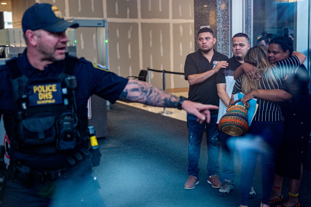 A DHS police officer gestures toward a family embracing at an ICE office.