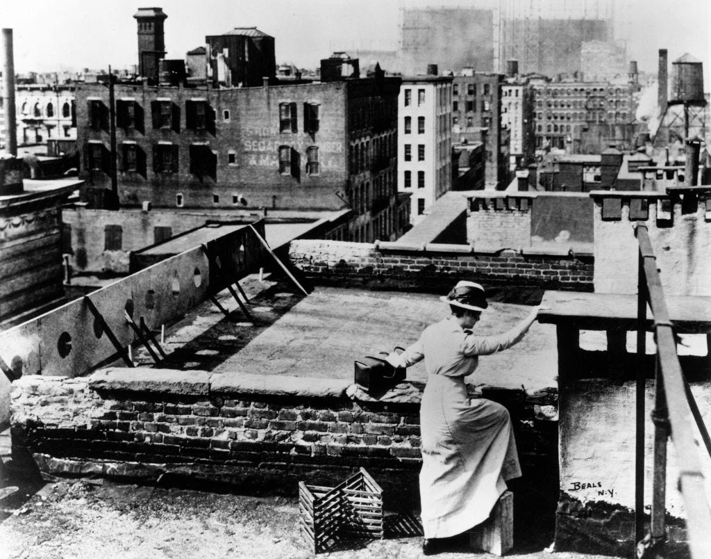 A lady is on top of a roof, trying to cross from one building to another. She is dressed in a long skirt, blouse, and hat.