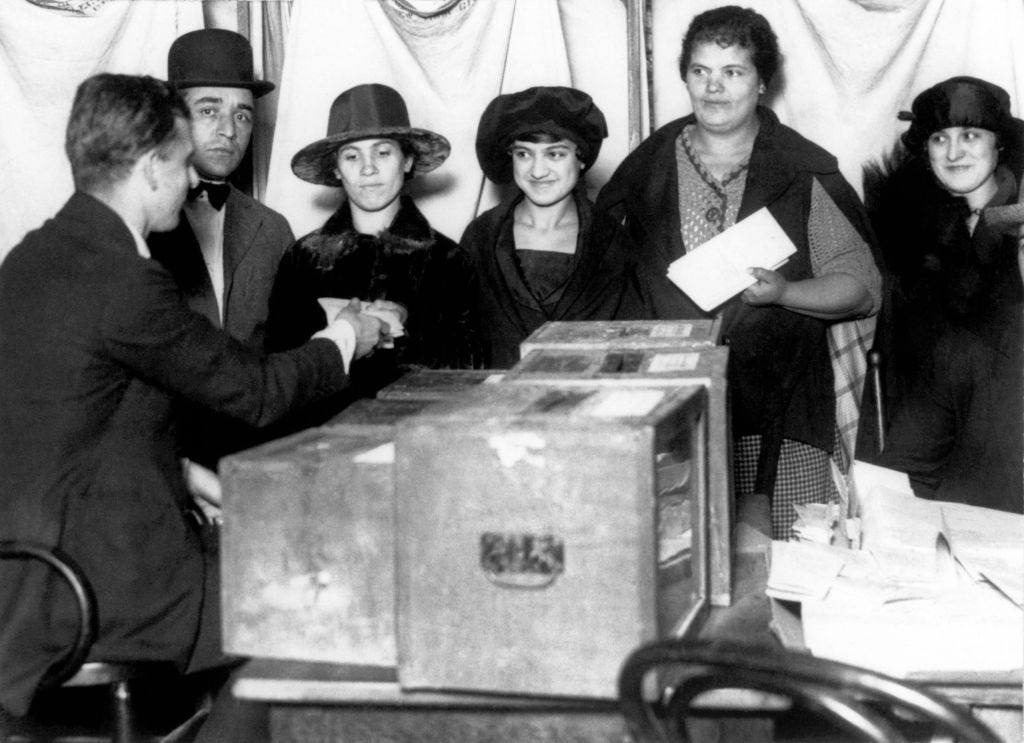 Two men are standing beside four women. One man is handing out a paper to one of the four women, who are all standing in front of a ballot box.
