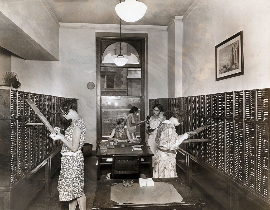 Five women in the office working on different tasks, from keeping records and checking papers to answering the phone.